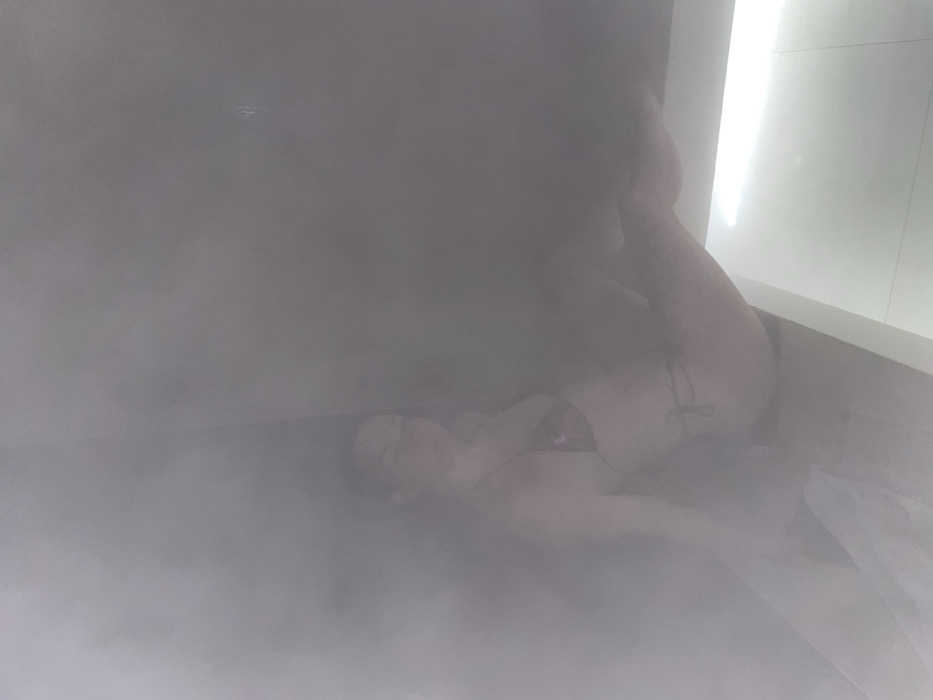 The image portrays a female subject in a densely steamy indoor environment, located in Boreti, Montenegro. A single woman, wearing a patterned two-piece swimsuit, is positioned in the left portion of the frame, seated or leaning against an indistinct dark surface. Her head is tilted slightly, and her limbs are flexed. The scene is enveloped in pervasive white steam or fog, significantly reducing visual clarity. In the lower right quadrant, a linear, bright artificial light source illuminates a light-colored, reflective surface, possibly a bench or floor. The upper walls of the room are dark and obscured by the steam.