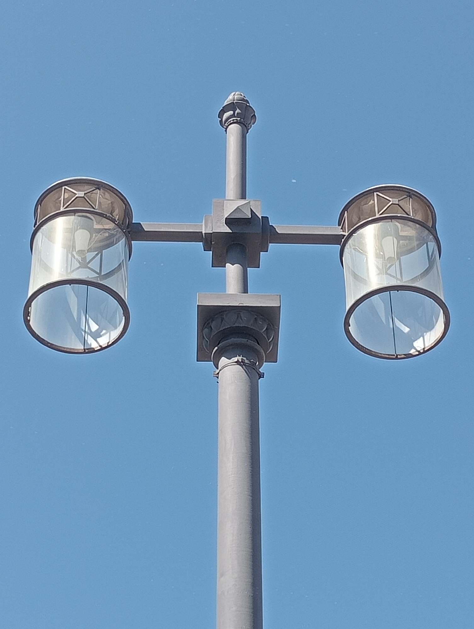 A gray lamppost with two lantern fixtures is shown against a clear blue sky. Each lantern has a clear cylindrical glass shade with a decorative metallic top. The lamppost has decorative detailing along its shaft. The image was taken in the city of Saint Petersburg, Russia.