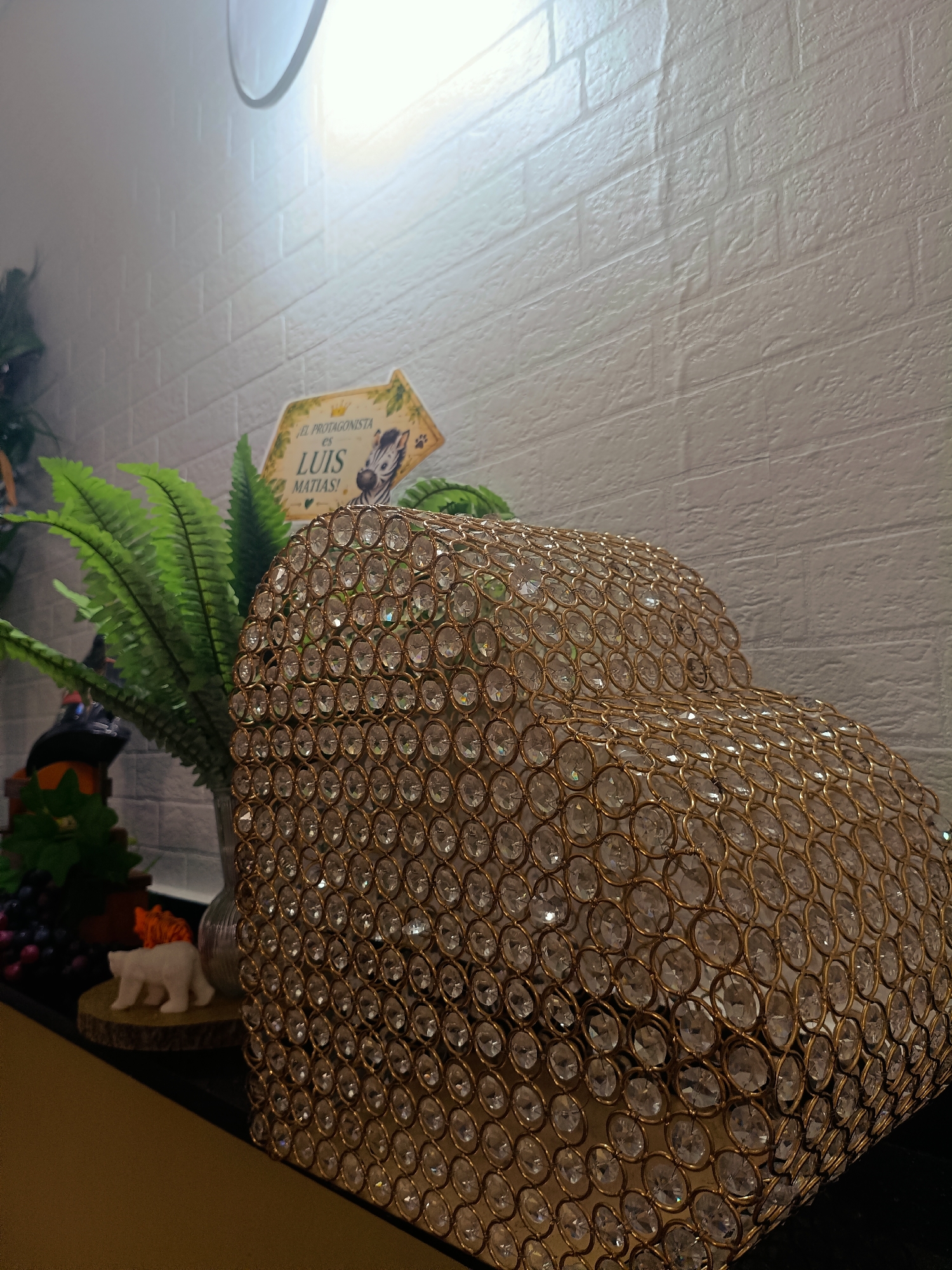 In the city of Rafelbunyol, Spain, a close-up shot captures a decorative arrangement on a surface, likely a shelf or table. The foreground is dominated by a large, heart-shaped object covered in gold-colored wire mesh and clear, faceted beads that reflect light. Behind this, a lush green fern-like plant is visible. Further back, a small, yellow, arrow-shaped sign with text and an image of a zebra is propped up. The background features a textured white brick wall. To the left, partially obscured, are elements of another decoration including what appears to be a small figurine of a white bear on a wooden disc, a cluster of dark grapes, and a Halloween-themed decoration with an orange pumpkin and a black witch hat. The lighting suggests an indoor setting.