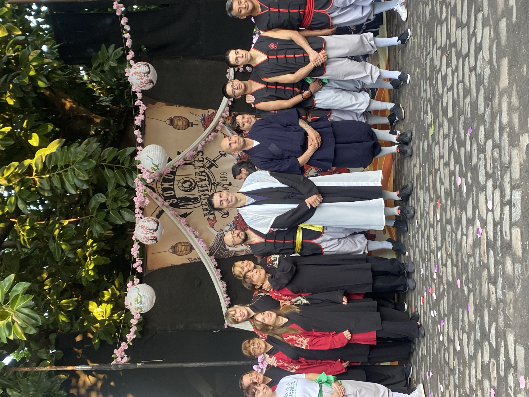 A group of approximately 13 people is standing in a line on a cobblestone street in what appears to be an outdoor setting. The individuals are dressed in martial arts attire, including black and red robes, black vests over gray pants, and one person in a white robe with a black trim. They are posing for a picture, possibly after a martial arts demonstration or event.
Behind the group, there's a decorated backdrop with a banner featuring the text "SOMOS MEDIEVALES ARG" and decorative lanterns hanging from a garland of pink flowers. To the left of the group, lush green foliage from trees is visible.
The location is likely within the city of Buenos Aires, Argentina, as indicated by the text on the banner, which translates to "We are Medieval Argentina." The event seems to be related to martial arts and medieval themes, taking place outdoors on a paved surface.