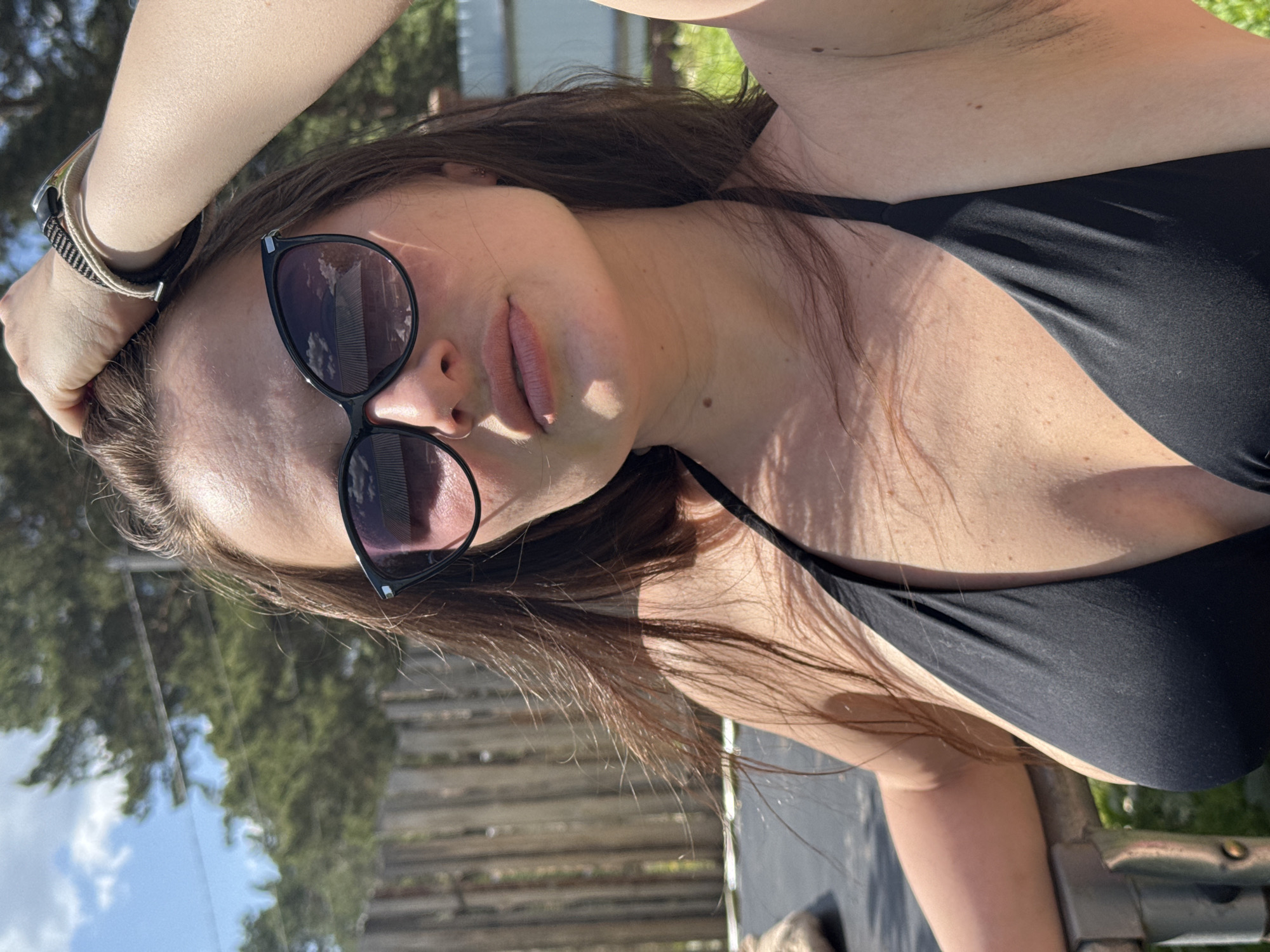 A woman wearing sunglasses and a black bikini top is posing for a photograph outdoors. She is fair-skinned with brown hair and has a watch on her wrist. The background includes trees and what appears to be wooden planks. It is not possible to confirm the location is Rusalevka, Russia.