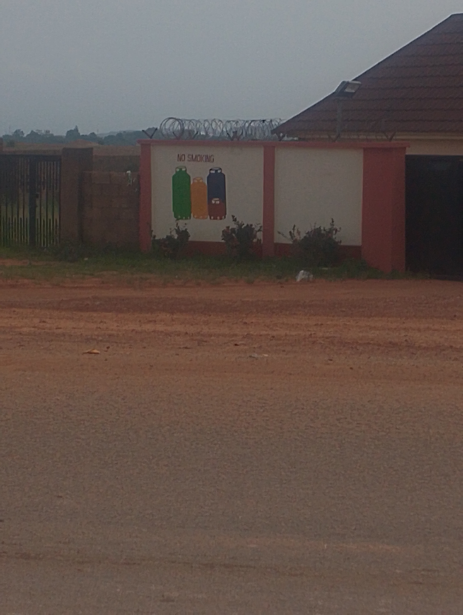 The image shows a section of a wall with a "NO SMOKING" sign and cartoon depictions of gas cylinders. The wall is painted white with red trim. To the left of the wall is a black metal gate and a brick fence. In the background, there is a building with a brown tiled roof and a light fixture attached to the roof. The area in front of the wall and fence appears to be an unpaved road or dirt area. The overall scene suggests an outdoor location, possibly a property boundary in a somewhat undeveloped area. The sky is overcast. There are no people or animals visible in the image. The location is specified as Jos, Nigeria, implying this is a scene from that city.