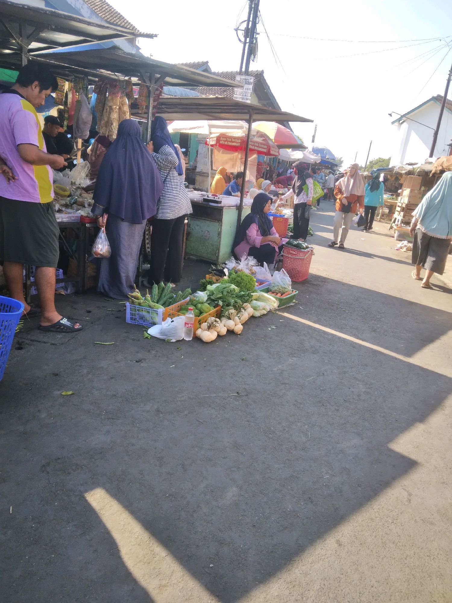 This is a photograph of an outdoor market scene in Sokaraja, Indonesia. The market appears to be set up along a paved road, with stalls constructed from wood and covered with awnings. The time of day is indicated by long shadows cast by the sun. In the foreground, a man wearing a purple and yellow shirt and dark shorts stands on the left, facing away from the camera. Next to him, a blue plastic basket is visible. A selection of vegetables, including cucumbers, lettuce, and some root vegetables, are displayed on the ground in crates and on plastic sheeting. A red woven basket with a cucumber inside is also present. A woman in a pink hijab and pink long-sleeved shirt is seated behind some of the vegetables, and appears to be interacting with a customer. Several other people, mostly women wearing hijabs of various colors, are visible in the background, walking through the market or attending to stalls. The stalls display a variety of goods, including packaged food items and more produce. The environment is typical of a bustling local market, with a mix of vendors, shoppers, and merchandise.