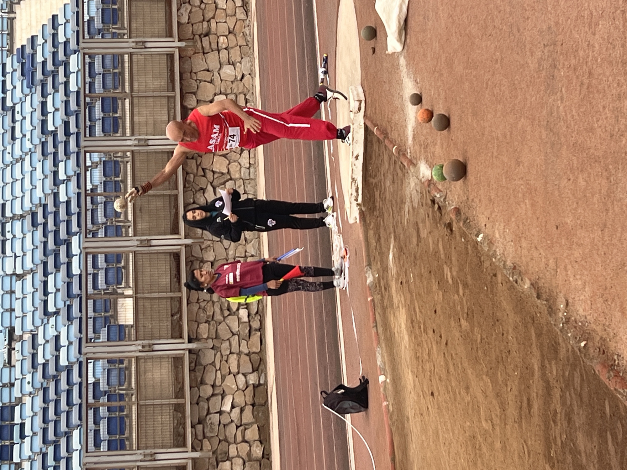 The image depicts a shot put event at the Regional Stadium of Arica, Chile. In the foreground, a male athlete wearing a red sleeveless shirt with "ASAM 74" printed on it, red pants with white stripes, and black socks with white and red shoes is in the process of throwing a shot put. He is on a concrete platform, with his right arm extended, holding the shot put. His body is angled, suggesting the culmination of his throwing motion.
Behind the athlete, two female figures are present. The one closer to the athlete is wearing a dark jacket with a white emblem on the chest and black pants, holding a paper and a pen. The other female, to the left, is wearing a maroon shirt with a yellow backpack and dark pants, holding a red horn and a blue object.
The background features a stadium with rows of blue seats. A stone wall runs along the base of the stadium seating. The ground is a reddish-brown dirt track, characteristic of athletic fields. Several shot put balls of varying colors, including green, brown, and orange, are scattered on the dirt surface in front of the throwing platform. A black backpack is visible on the edge of the track to the left of the frame, connected by a white cord. The overall scene captures a moment of athletic competition in an outdoor stadium.