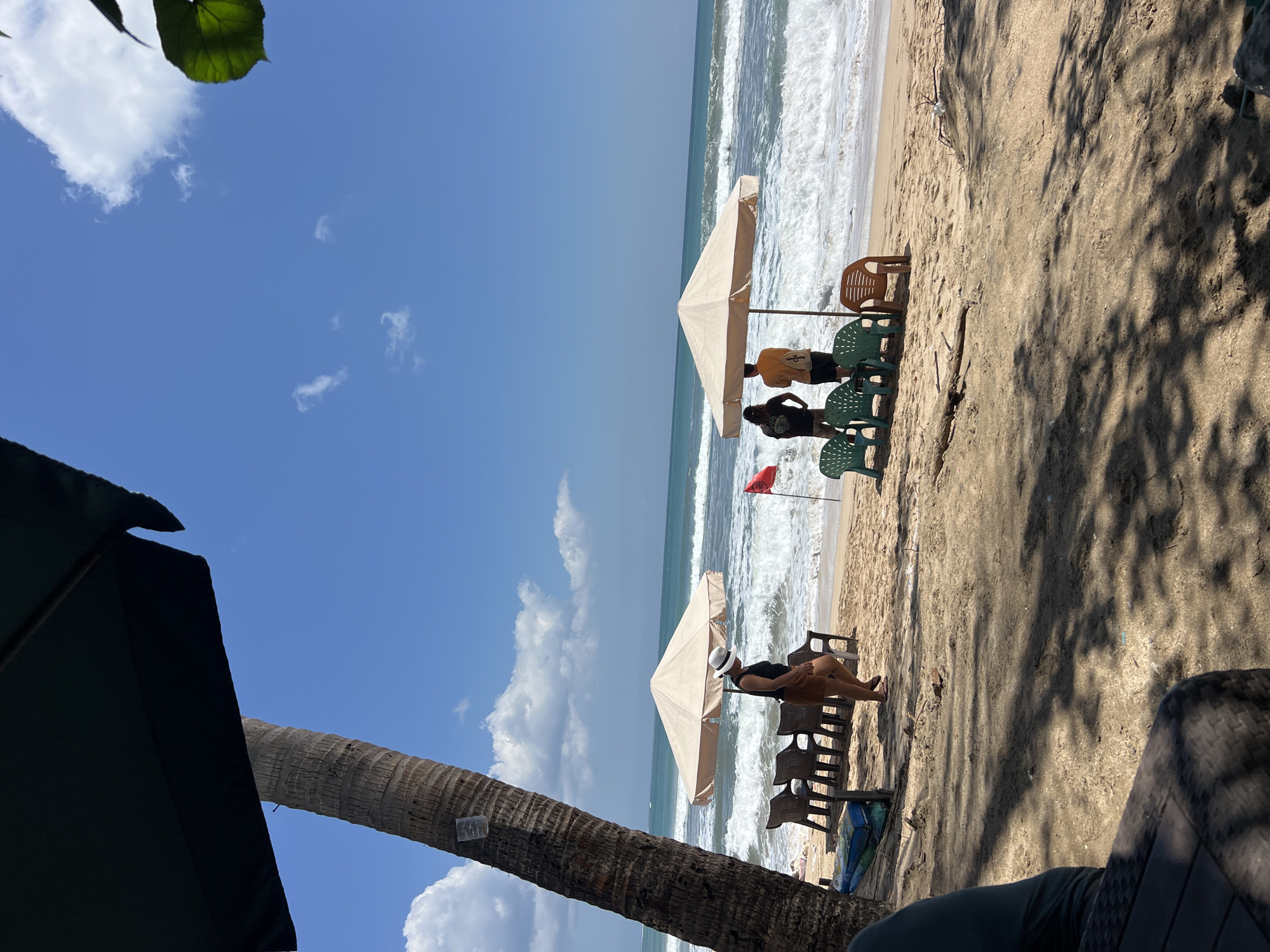 A low-angle, long shot captures a sandy beach in Kuta, Indonesia, with a palm tree on the left and a large dark umbrella in the foreground casting shadows. The ocean is visible with moderate waves and white foam breaking on the shore. Two beige beach umbrellas stand on the sand, each with several plastic chairs arranged around them. In the middle ground, a woman wearing a white hat and brown shorts walks near one umbrella, while two people, one in a black shirt and the other in a yellow shirt, stand near the second umbrella. A red flag is planted in the sand between the umbrellas. The sky is blue with scattered white clouds.
