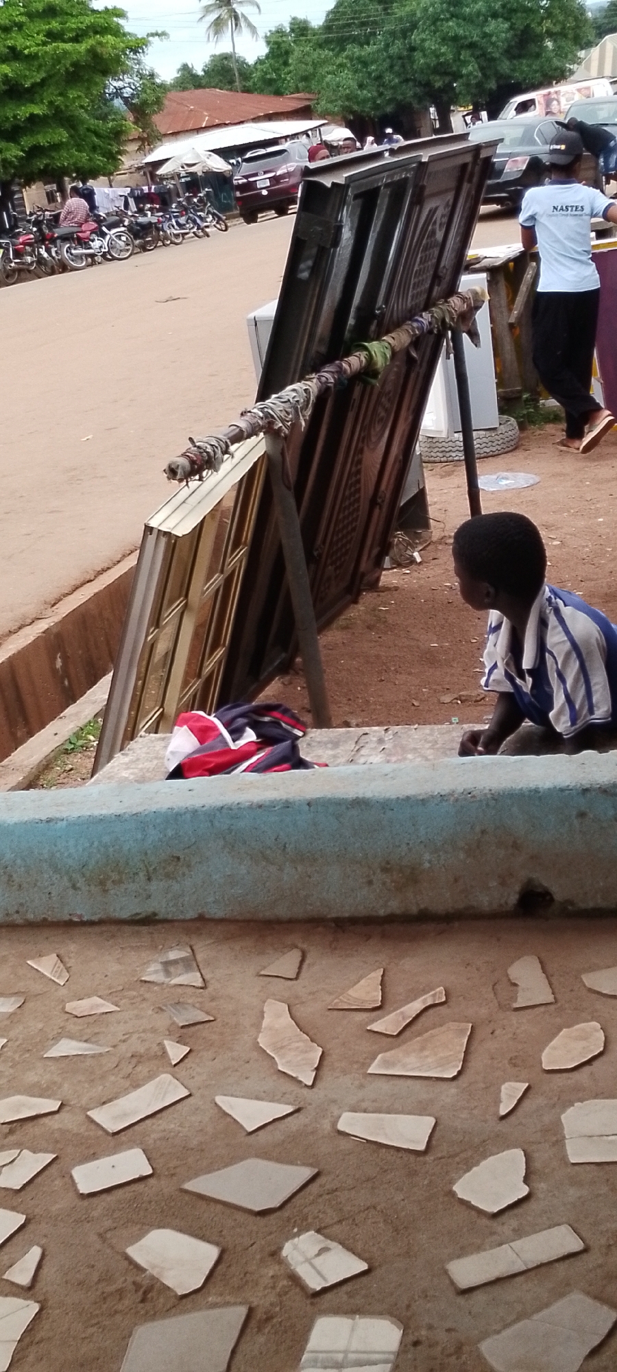 In Gboko, Nigeria, an outdoor scene unfolds with a focus on a young boy sitting on a concrete step, looking towards a display of metal doors. The doors, leaning against a metal stand wrapped with colorful fabric, are ornately decorated and appear to be for sale. In the background, a red SUV is parked on a dirt road, alongside a row of motorcycles. Further back, buildings with corrugated iron roofs suggest a bustling market or roadside settlement. A woman in a light blue shirt with "NASTES" printed on it walks away from the camera, while a few other individuals are visible in the distance. The ground in the foreground is covered with scattered, irregular pieces of broken tiles, forming a mosaic-like pattern on the dusty earth.