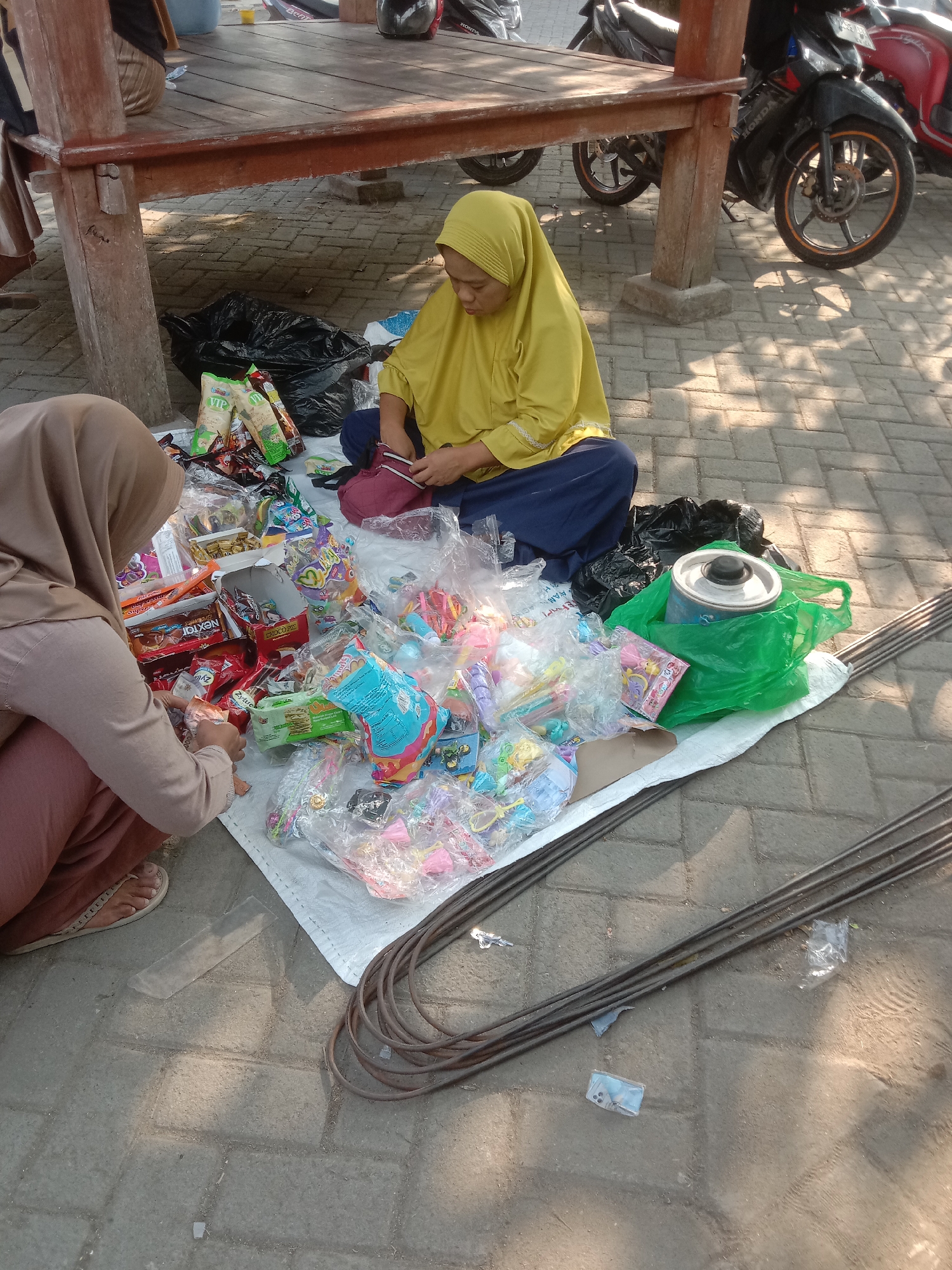 Two women are seen squatting on a paved ground in an outdoor setting, possibly a market. The woman on the left is wearing a light brown hijab and a matching colored skirt, and is focused on arranging small items in plastic packaging. The woman on the right is wearing a bright yellow hijab and a dark blue skirt, and is in the process of sorting through a pile of colorful items, some of which appear to be toys or candy. Both women are surrounded by various goods laid out on a white cloth, including bags of snacks, small toys, and other packaged items. A large black trash bag and a green plastic bag containing a metal container are also visible. The ground is paved with interlocking bricks. In the background, a wooden structure resembling a stall or seating area is present, and motorcycles are parked nearby, suggesting an urban or semi-urban environment. The scene is illuminated by sunlight, creating shadows on the ground. The overall impression is one of commerce or preparation for sale.