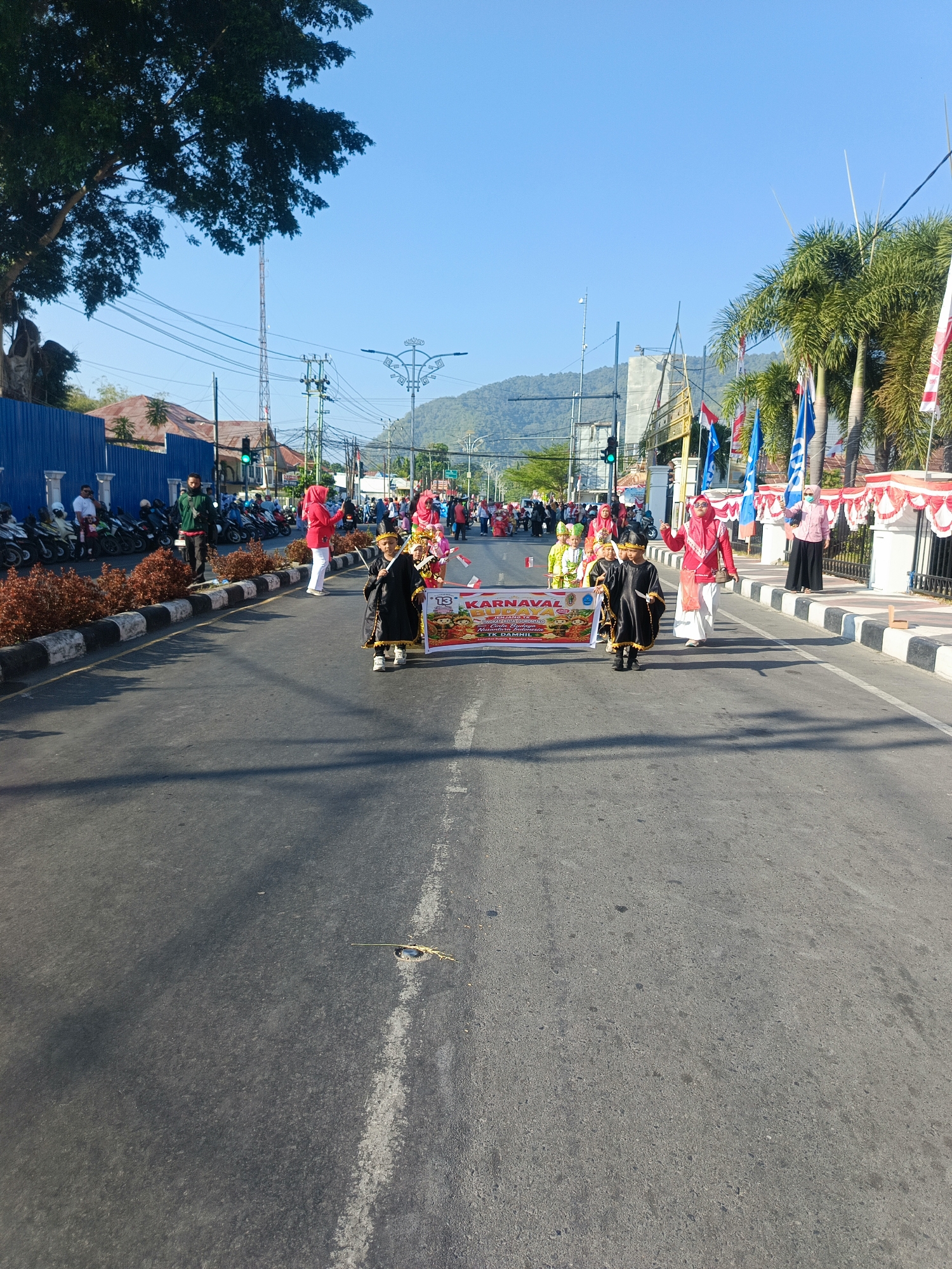 A parade is taking place on a paved road in Gorontalo, Indonesia, under a clear blue sky. In the foreground, two children dressed in traditional black and gold attire are walking, holding a banner that reads "KARNAVAL BUDAYA" and depicts cartoon characters. Several other children in colorful costumes, along with adults in festive clothing, are participating in the parade further down the street. The road is lined with motorcycles on the left side and decorative red and white banners and palm trees on the right. Buildings and a distant mountain range are visible in the background. The overall scene depicts a cultural celebration.