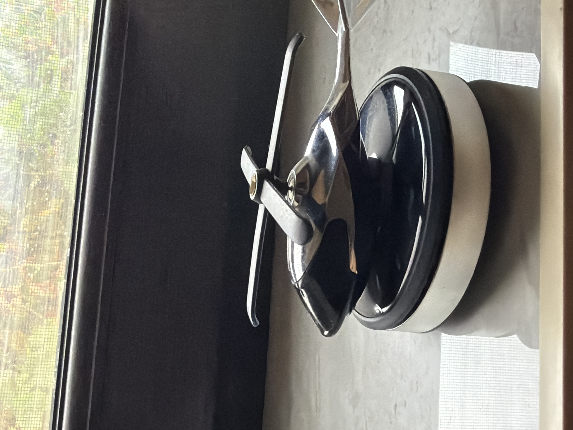 The image displays a chrome-plated model helicopter resting on a black and silver base. The helicopter has a sleek, modern design with two visible rotors. The base appears to be made of plastic with a metallic accent. The scene is set on a reflective surface, possibly a countertop or table, which casts a subtle shadow of the helicopter. In the background, a window is visible with a screen, suggesting an indoor setting. The view through the window shows blurred greenery, possibly indicating a garden or natural landscape. The overall lighting is soft, with highlights reflecting off the polished surfaces of the helicopter and base. The image does not provide any information or context related to the city of Sabirabad, Azerbaijan.