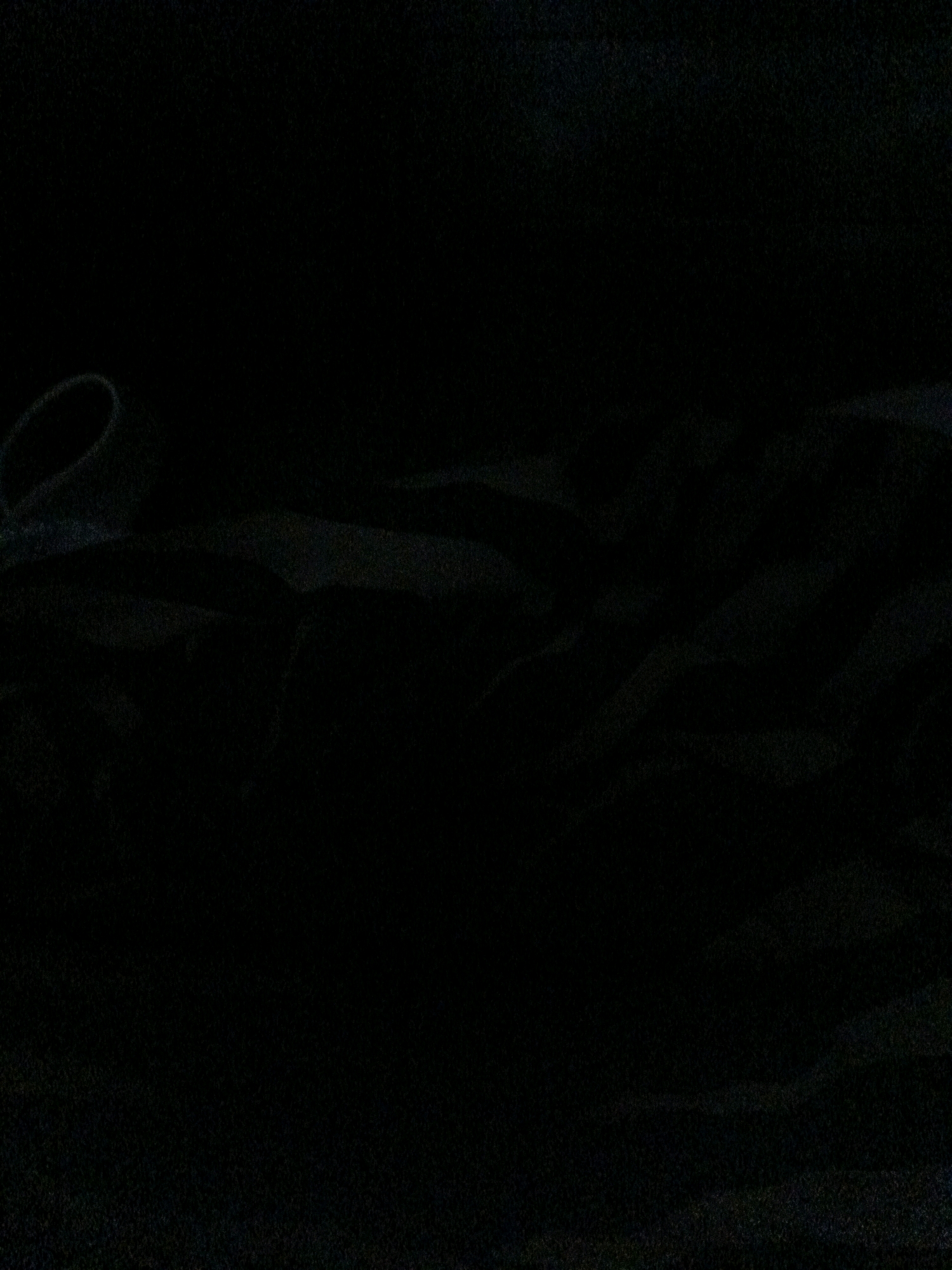 The image is a dark, low-light photograph of a black fabric. The fabric appears to be textured and possibly crumpled or folded, creating subtle shadows and highlights. In the upper left portion of the image, a curved edge or loop of the fabric is visible, outlined by a faint, lighter tone, suggesting a material like nylon or a synthetic blend. The rest of the image is dominated by deep shadows with only very faint hints of lighter areas suggesting folds or creases in the fabric. There are no discernible objects, people, actions, interactions, or environmental details visible in the scene. The photograph provides no information regarding its location, including West Jakarta, Indonesia.