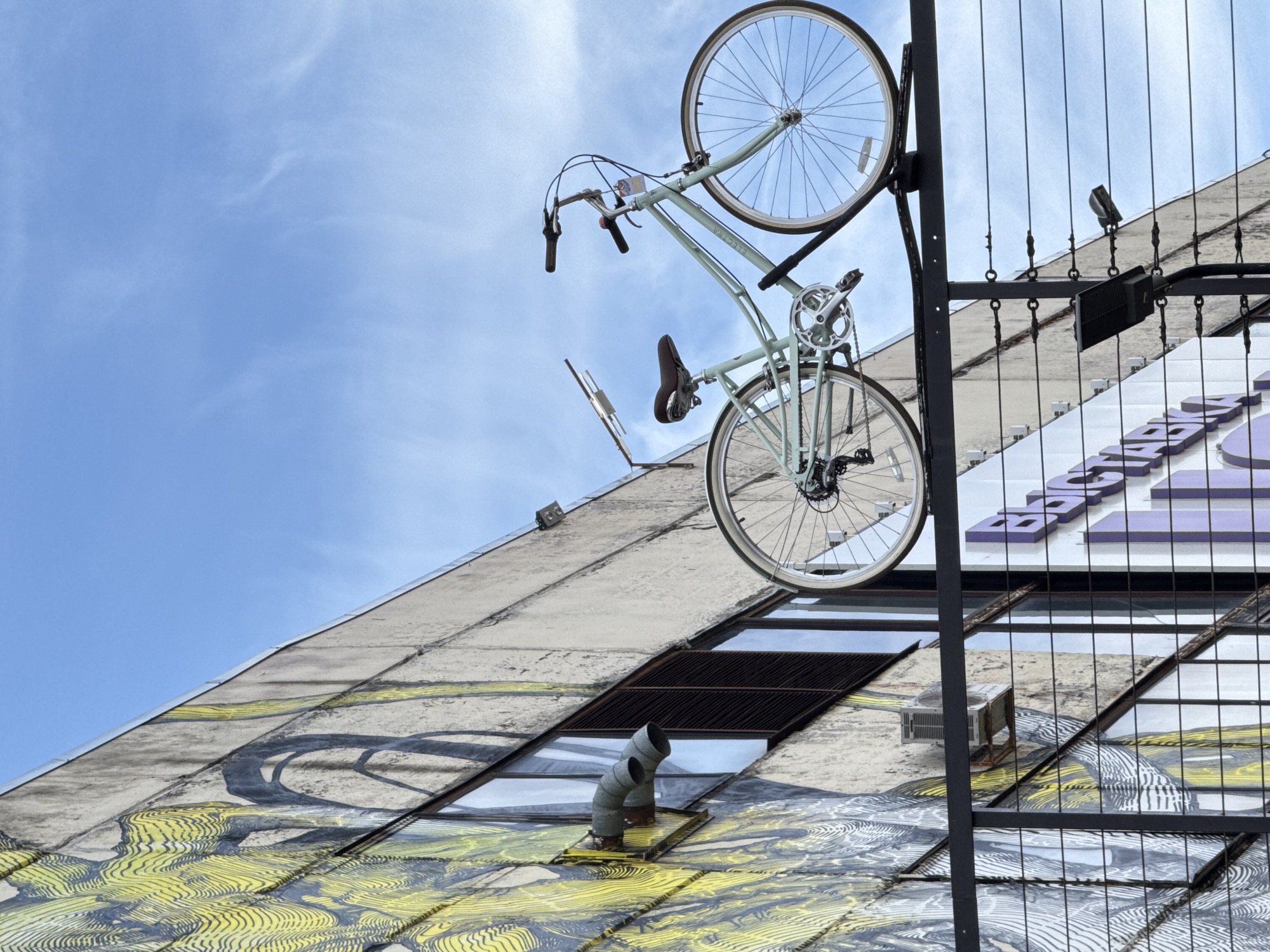 A low-angle view shows a light blue Electra bicycle suspended from a black metal structure against a partly cloudy blue sky and a building façade. The bicycle is oriented upside down, with its wheels pointing upwards. It has white wheels, black tires, and a brown seat.
The building's facade is a mix of painted design (yellow and black) and textured concrete. A ventilation pipe protrudes from the building. Parts of a sign with purple lettering are also visible on the building's facade.
The black metal structure that supports the bike appears to be part of a fence or decorative element attached to the building. Its vertical lines intersect with the building's angles, creating a geometric composition.