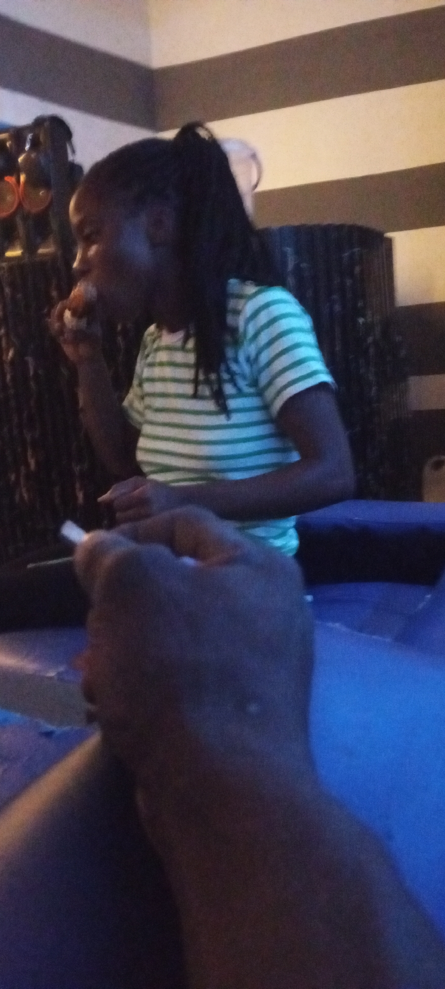 In Adankolo, Nigeria, a person is seen in a dimly lit indoor environment, possibly a lounge or entertainment area. The person, with dark braided hair pulled into a ponytail, is wearing a green and white horizontally striped t-shirt. They are holding a small, light-colored object, possibly food, up to their mouth, indicating they are in the process of eating. Another person's hand is visible in the foreground, holding a cigarette. The background features a wall with alternating dark and light horizontal stripes and dark vertical blinds or curtains. The overall atmosphere suggests a casual gathering or personal moment.