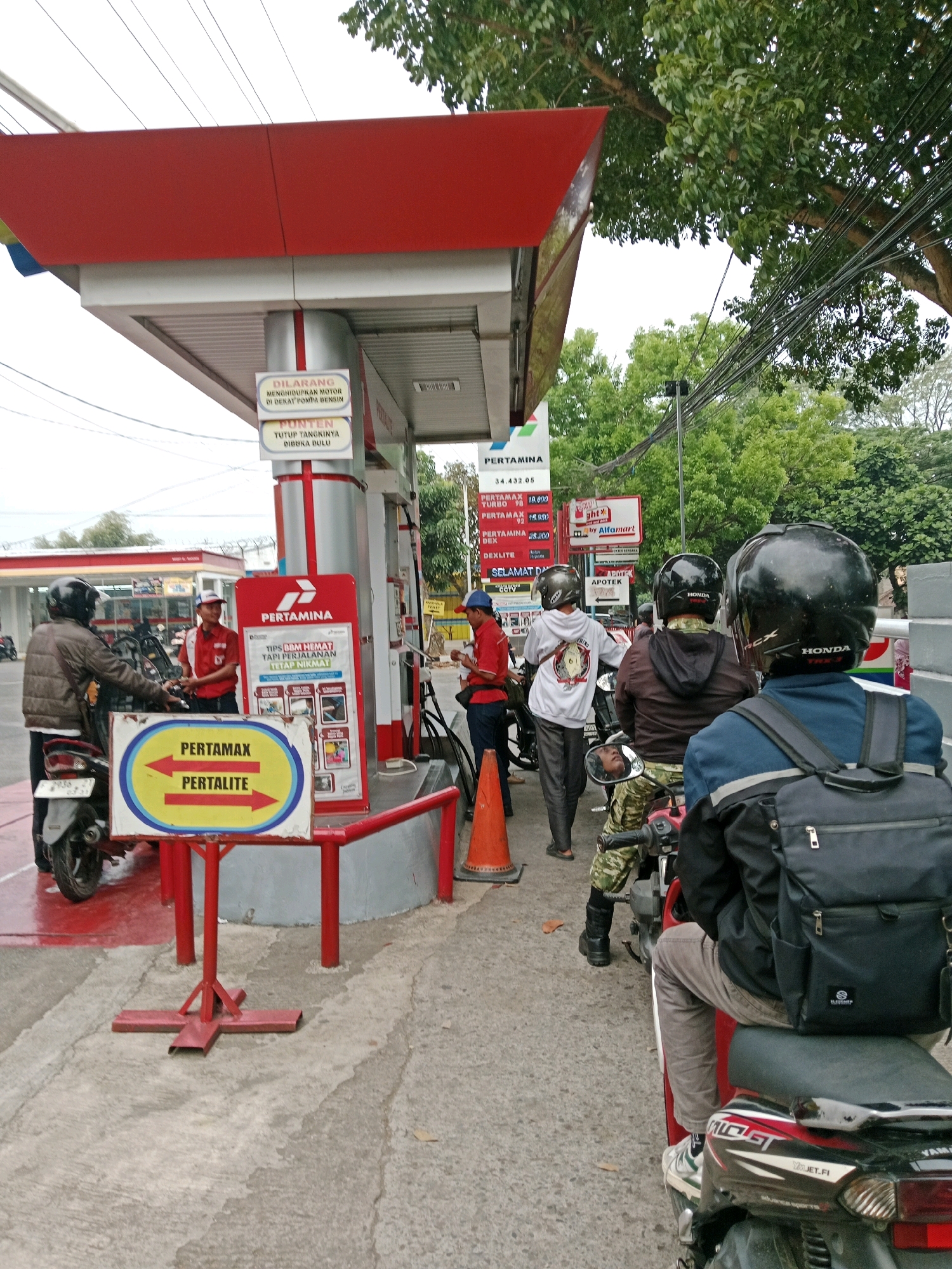 The image depicts a Pertamina gas station in Muka, Indonesia, with several people on motorcycles queuing to refuel. In the foreground, a person wearing a black jacket and helmet is seated on a red motorcycle, facing away from the camera. To their left, another person in a brown jacket and helmet is on a motorcycle, speaking with a gas station attendant wearing a red uniform. In the background, several other motorcycles and individuals are visible, some receiving service from attendants.
The gas station has a red and white canopy with Pertamina branding. Signs display fuel prices and promotions. The environment appears to be an urban or suburban setting, with trees and power lines visible in the background. The ground is paved, and there are some fallen leaves. An orange traffic cone is placed near one of the fuel pumps. The overall scene suggests a typical day at a busy gas station.