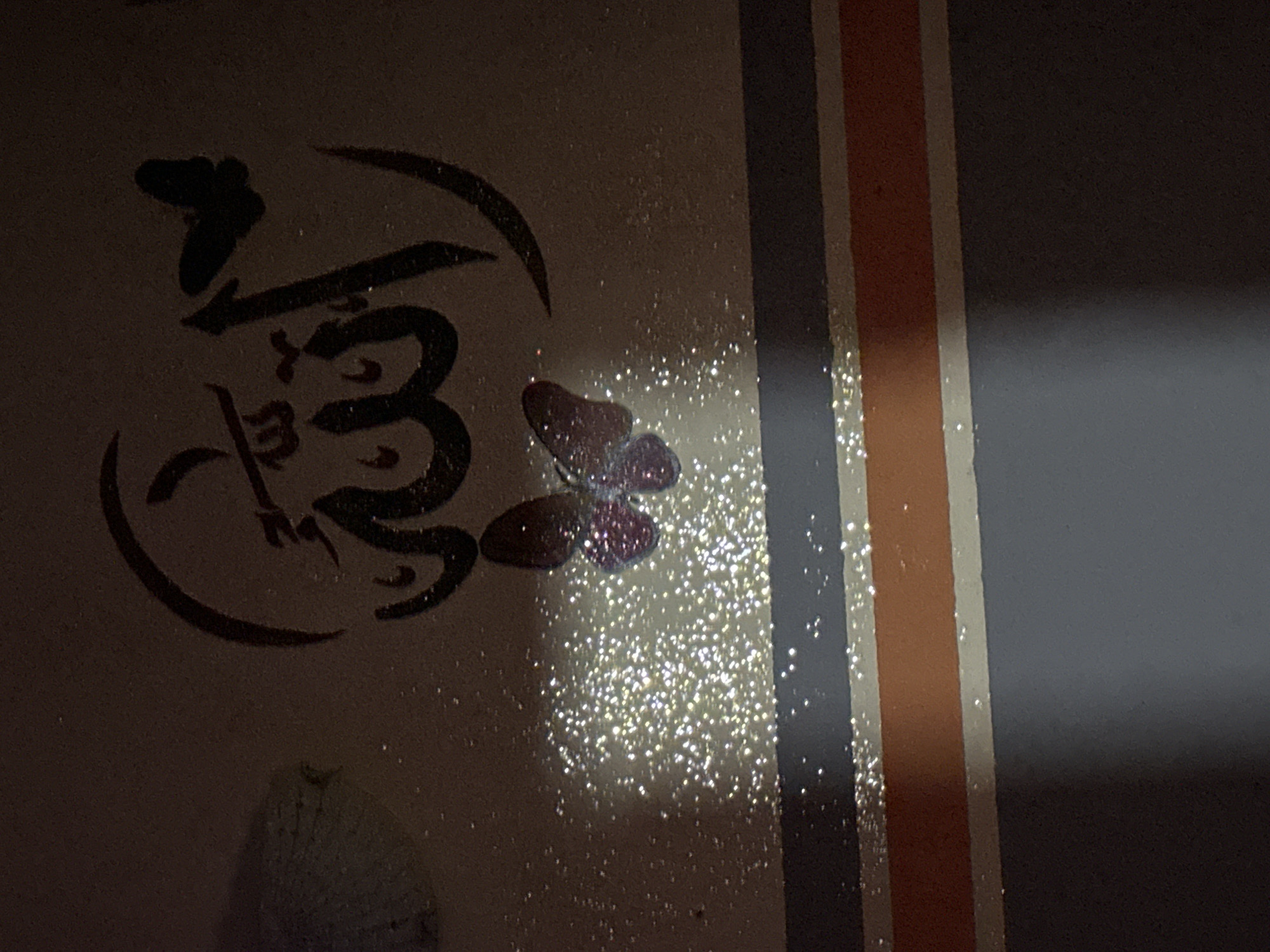 A close-up shot shows a section of a surface, potentially a wall or board, featuring artwork and design elements. On the left, there's dark, calligraphic-style lettering enclosed within a curving shape. To the right of the calligraphy is a butterfly-shaped object with a dark body. The area around the butterfly and below the calligraphy is heavily scattered with what appears to be glittering particles, possibly glitter or shiny dust, catching the light. On the right side of the image, there are vertical stripes in shades of orange, white, and gray. The gray stripe appears to extend into a darker, shadowed area on the far right of the frame. The overall lighting is somewhat dim.