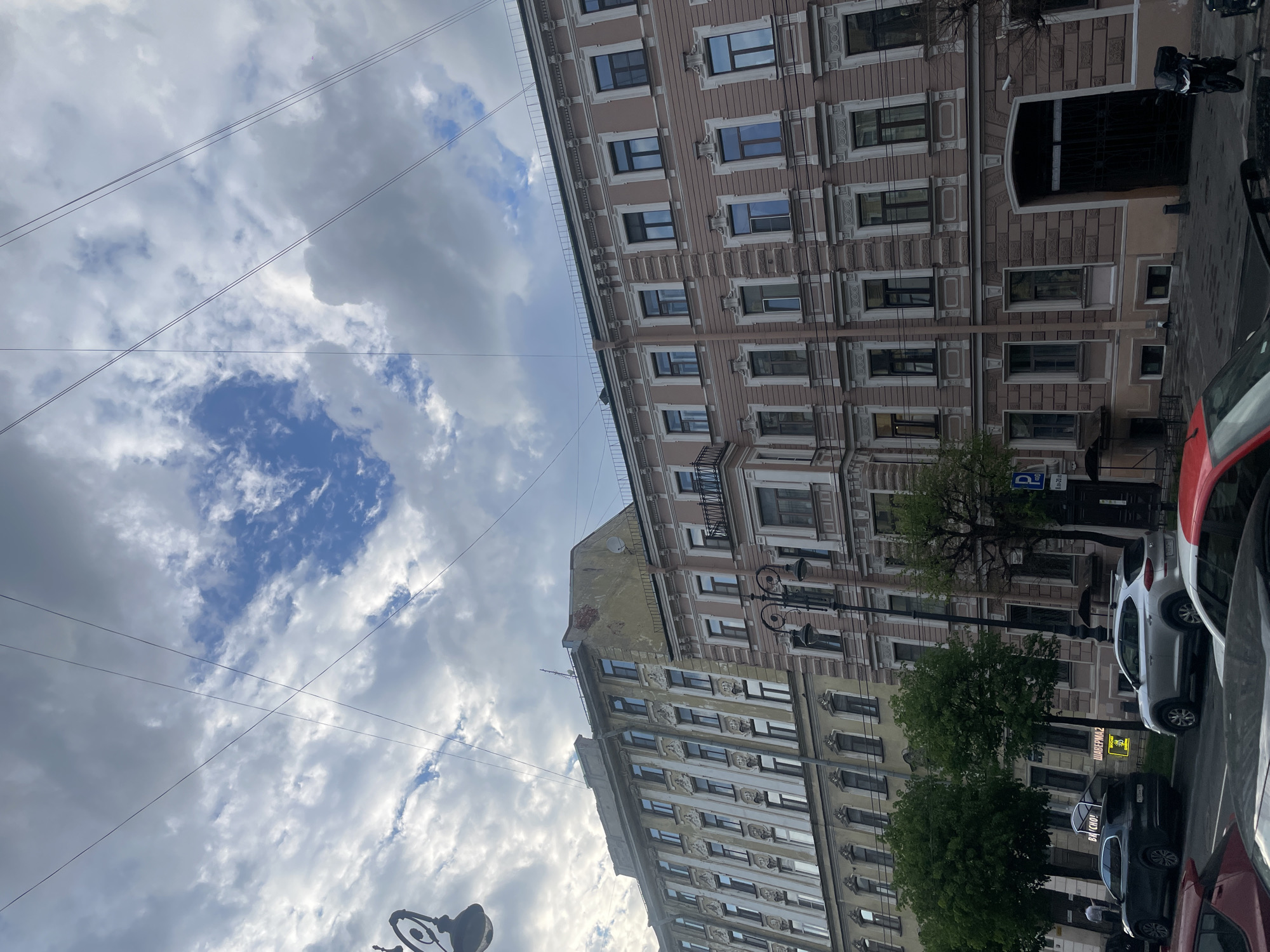 The image, oriented vertically, captures a bustling street scene in Saint Petersburg, Russia, under a partly cloudy sky. Dominating the frame are two ornate, multi-story buildings, characteristic of the city's architecture. The building on the right is a pale pinkish-beige, featuring numerous windows with decorative frames, cornices, and a small balcony on one of the lower floors. The building on the left is a yellowish-beige, with similar architectural detailing, though its upper section shows some signs of wear and peeling plaster. A satellite dish is visible on the roof of this building.
Several utility wires crisscross the sky, intersecting near the center of the frame against patches of clear blue sky peeking through the clouds. Along the bottom and right side of the image, a street is visible with multiple cars parked parallel to the curb, including a red car, a white SUV, and a dark-colored car. Ornate streetlights stand at intervals along the street. Green foliage from several trees is scattered in front of the buildings, obscuring parts of the lower floors.
Notable details include a blue parking sign displaying "P" and "8-20" (likely indicating hours of operation) and a glowing yellow-green sign advertising "ШАВЕРМА" (Shawarma), with "ВЫХОД" (Exit) also faintly visible. On the rear window of the black car, the text "БА ВКУСНО!" (suggesting "delicious!") can be read. A motorcycle or scooter is parked near a large, arched entrance to one of the buildings. The time of day appears to be daytime, with ample natural light.