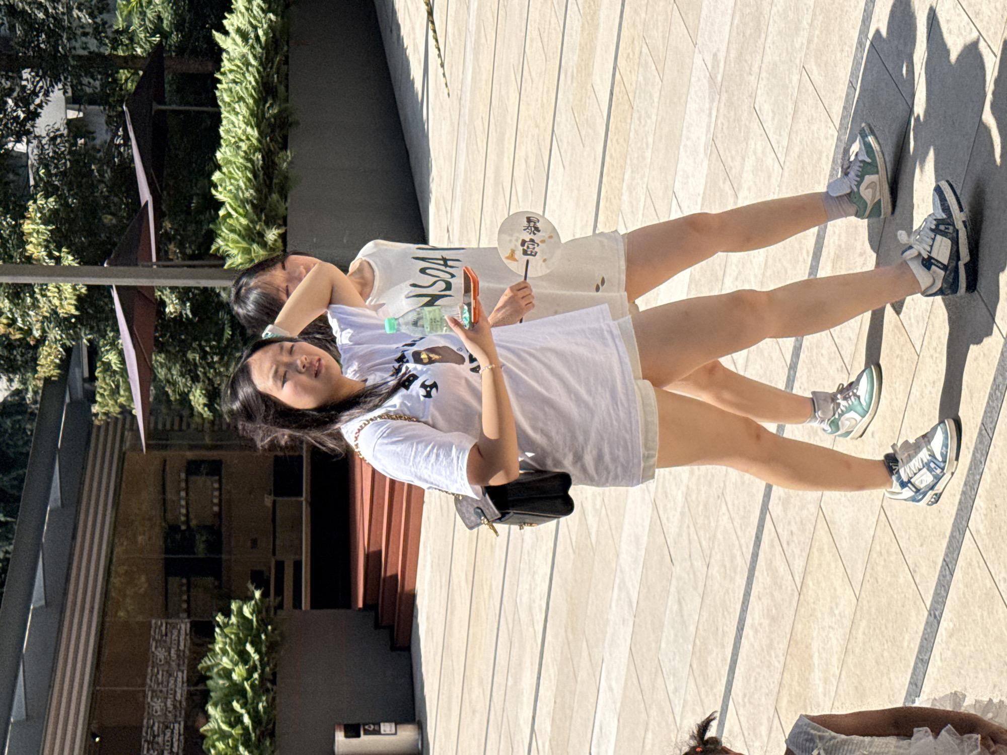In Yuzhong District, China, two young women walk along a paved outdoor area bathed in sunlight. The woman in front, wearing a white t-shirt and shorts, carries a black handbag over her left shoulder and holds a smartphone and a bottle of water. She is also holding a small fan in her right hand, which is partially obscured by her arm. The woman behind her, also in a white t-shirt and shorts, has her right arm raised, touching her head. A young child with pigtails stands to the far left of the frame, facing away from the camera. In the background, there is a building with large glass windows and outdoor umbrellas. The ground is a light-colored, tiled surface with distinct grout lines.