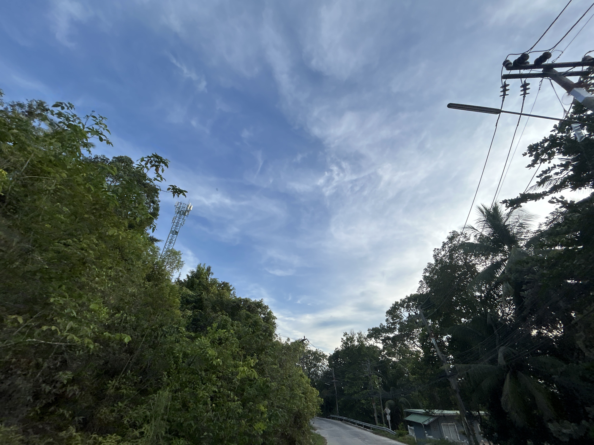 The image depicts a road in Baan Wang Ta Kien, Thailand, bordered by lush vegetation on both sides. A tall telecommunications tower is visible through the trees on the left. Power lines and a street light are on the right. The sky is blue with scattered clouds. A building is nestled in the trees on the right side of the road.