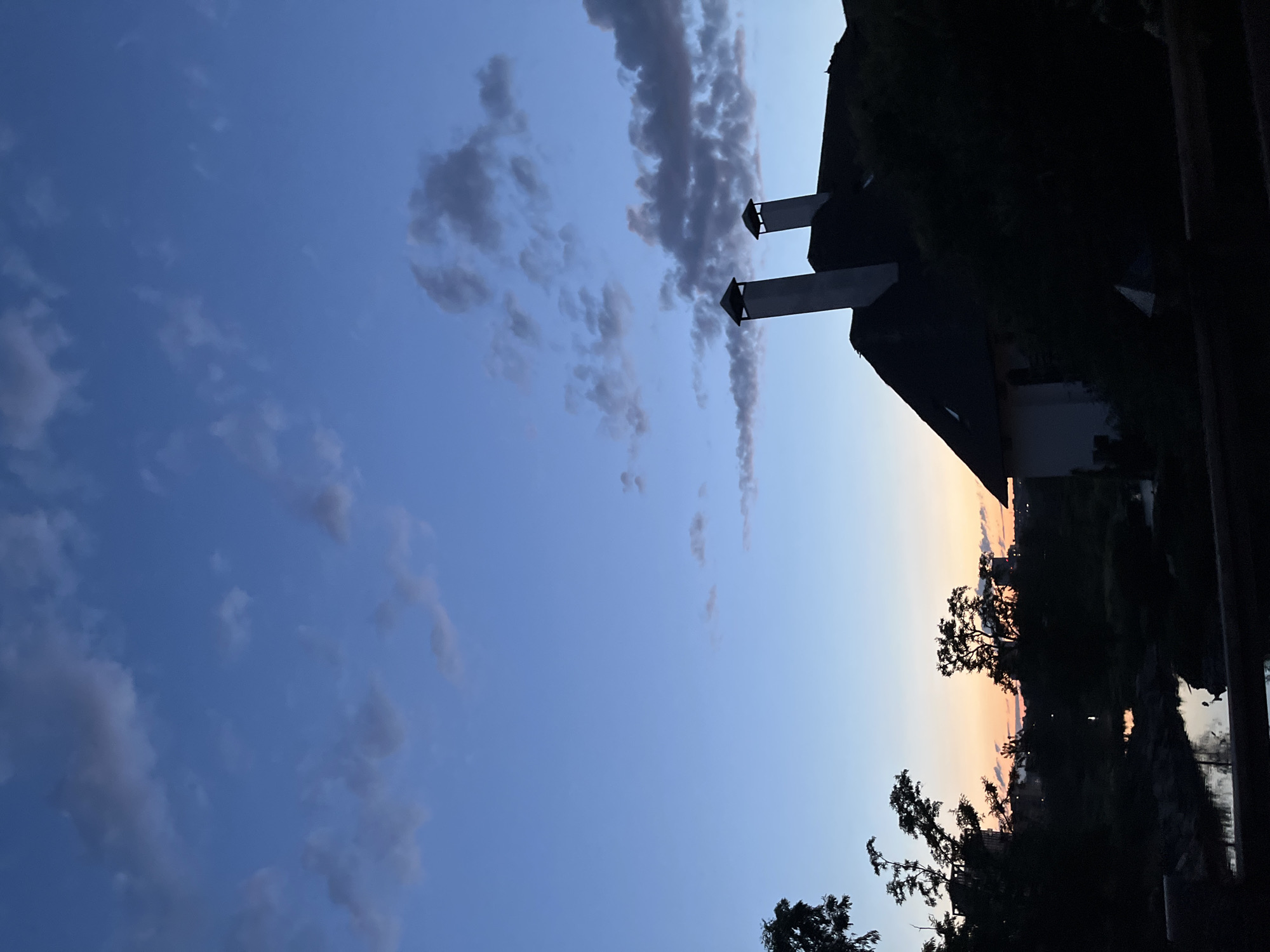 The image depicts a tranquil evening scene in the city of Dnipro, Ukraine, likely during dusk. The sky is a gradient of deep blue at the top, transitioning to lighter shades of blue and a soft orange hue near the horizon, indicating the last light of the day. Scattered clouds, tinged with grey and pink, drift across the sky.
Dominating the right side of the frame is the silhouette of a large house with a dark, sloped roof. Two prominent, rectangular chimneys rise from the roof, their tops capped. The house appears to be partially obscured by dense, dark foliage in the foreground.
On the left side of the image, silhouetted trees with irregular branches frame the scene. In the middle ground, a body of water, possibly a river or a canal, reflects the colors of the sky. The water's surface is calm, with subtle ripples. Beyond the water, more trees and some faint structures of buildings are visible in the distance, softened by the twilight. A dark railing runs along the bottom of the image, suggesting a viewing platform or a fence. The overall atmosphere is one of quiet and natural beauty.