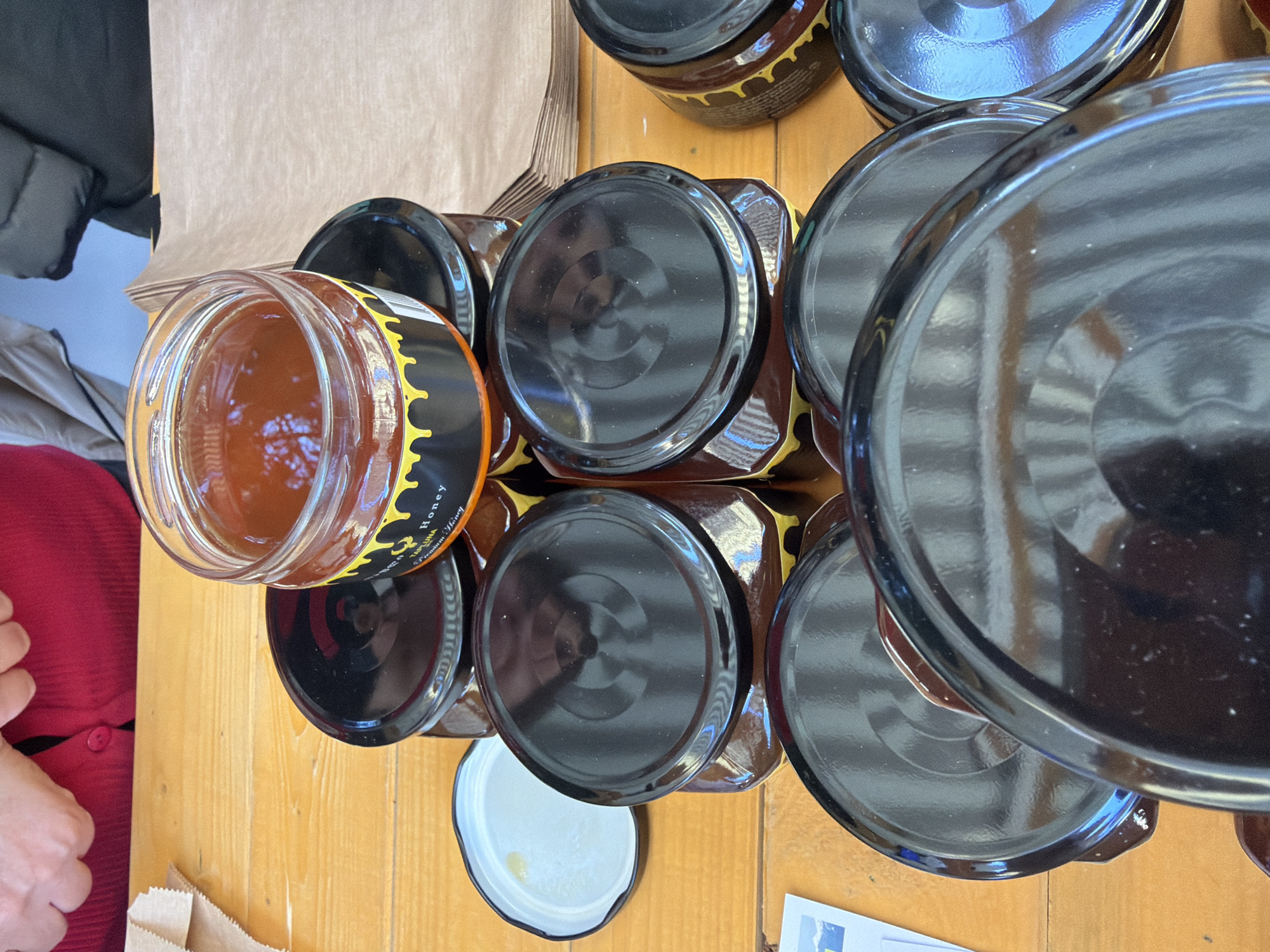 Overhead shot of jars of honey on a wooden table. There are at least eight jars, most with black lids. One jar is open, revealing amber-colored honey. The jar has a black label with yellow dripping design. Some jars have brown wrapping around the sides. The table is outdoors, possibly at a market. A person wearing a red sweater is visible on the left. Paper bags are also on the table. The city of Tbilisi, Georgia, is not evident.