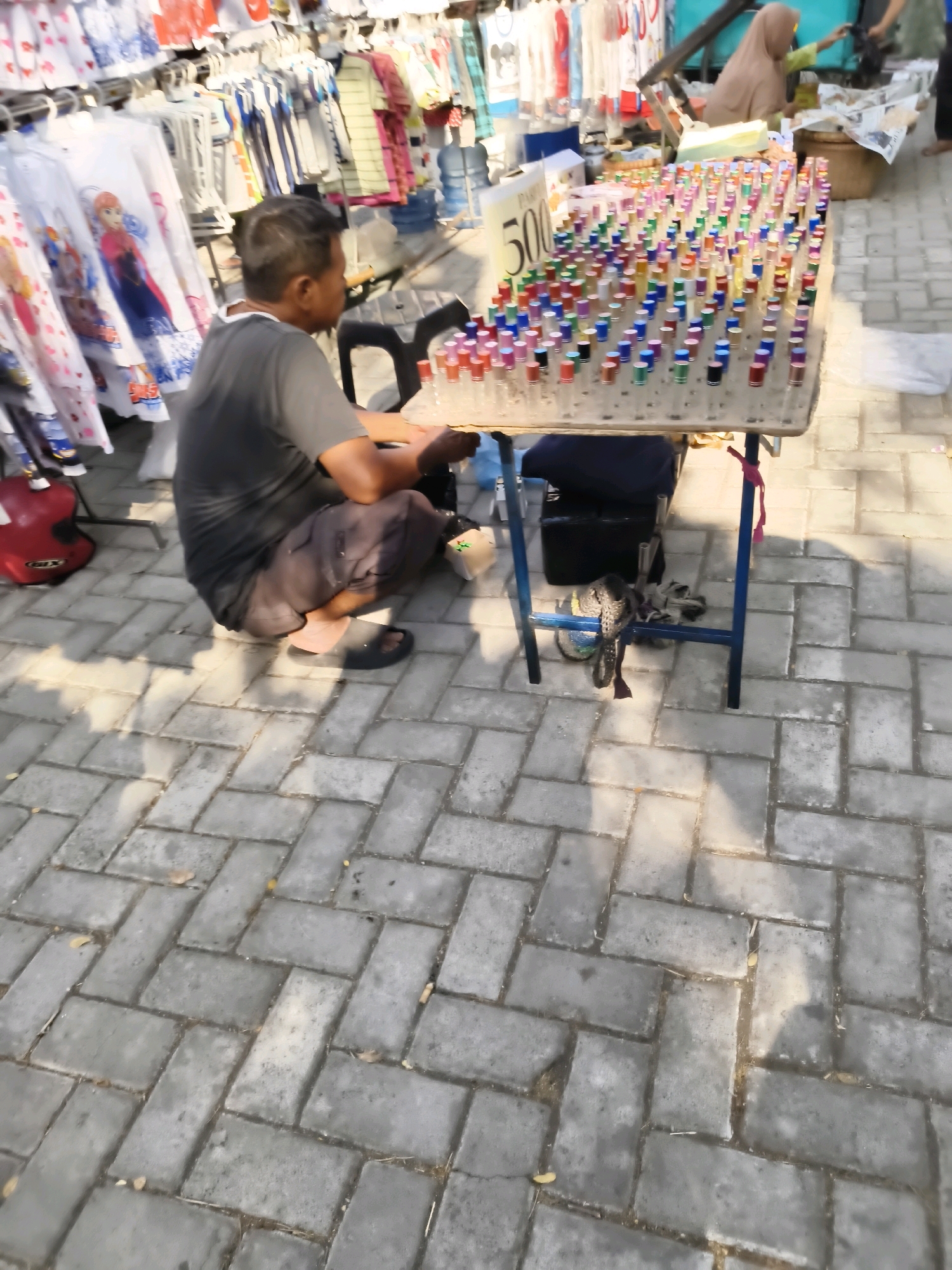 The image depicts a street vendor in Kedungan, Indonesia, selling small bottles of perfume from a portable table. The vendor, a man in a dark t-shirt and shorts, is crouched beside the table, his face turned away from the camera. The table is laden with rows of colorful perfume bottles with various cap colors, suggesting a wide selection. Behind the vendor, a rack of clothing, possibly for sale as well, is visible. A red helmet rests on the ground near the vendor. In the background, another person, partially obscured by a hijab, appears to be interacting with something out of frame, and a shopping basket is also present. The scene is set on a paved walkway, with sunlight casting long shadows, indicating it is daytime.