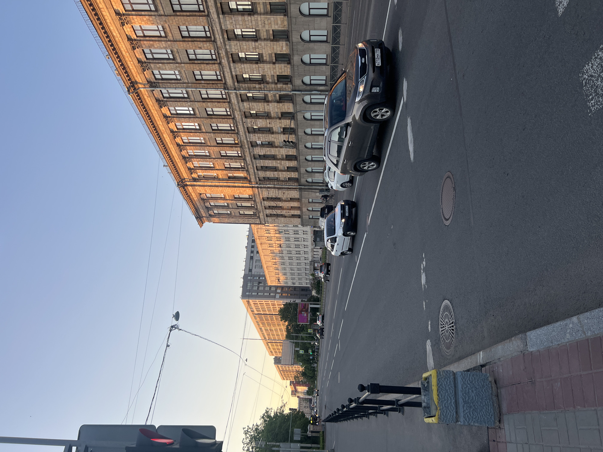 This forensic description details an urban street scene in Saint Petersburg, Russia, captured during the late afternoon or early evening under low, golden sunlight.
The image presents a wide, multi-lane asphalt road (at least four visible lanes) with a median, extending into the distance. White dashed and solid lines mark the lanes. On the right side of the road, a grand, multi-story building with classical architectural elements stands prominently. It features numerous rectangular windows, stone pilasters between sections, and a distinctive cornice, all brightly illuminated by the setting sun, giving its facade a warm, golden hue.
On the left side of the street, a paved sidewalk with reddish-brown and grey pavers is bordered by a black metal fence with decorative finials. A grey concrete trash receptacle with a yellow rim is situated on the sidewalk in the lower left foreground. A traffic light pole is visible on the far left, with its signal displaying red. Above the road, utility wires and streetlights are suspended.
Further down the street on the left, a series of other multi-story buildings are visible, some appearing modern with grey facades, while others exhibit older architectural styles. These buildings also catch the golden light of the sun. Trees line the left side of the road between the sidewalk and the buildings.
Vehicles are present on the road, all moving in the direction away from the viewer. In the mid-ground, a dark grey Kia Sorento SUV (license plate A713MH 198) occupies the second lane from the right. Behind it, a white car follows. To the left of the Kia, in the leftmost visible lane, a white sedan (license plate K086OE 81) is driving. Additional vehicles are visible further down the road.
The sky overhead is a clear light blue, transitioning to a pale yellow-orange gradient near the horizon, indicative of a sunset or sunrise. Manhole covers and storm drains are visible on the road surface. No discernible people are prominently featured in the immediate foreground or mid-ground.