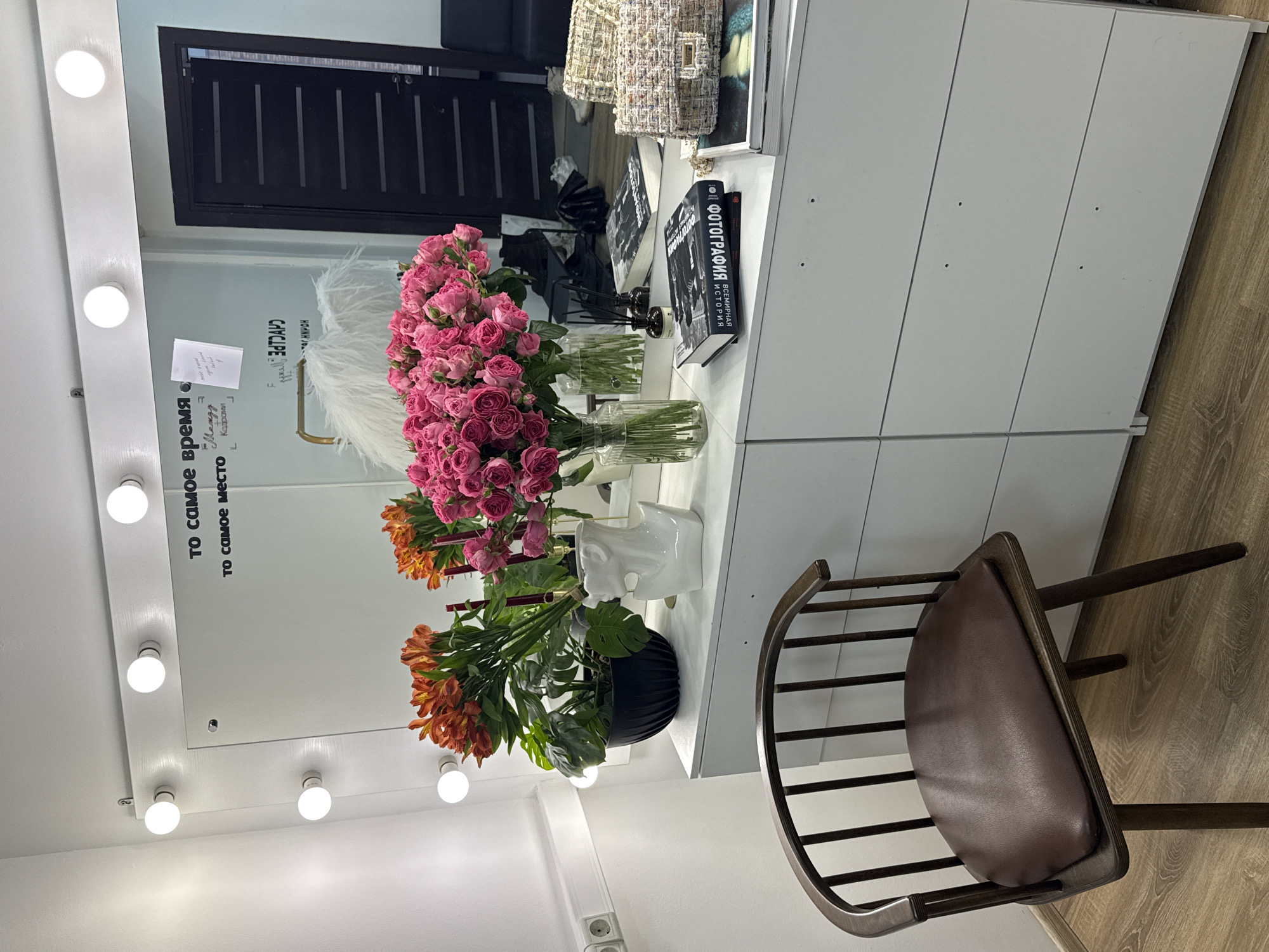 The image shows an interior scene with a makeup vanity positioned against a wall. The vanity is white and features a mirror surrounded by light bulbs. Reflected in the mirror are various objects, including bouquets of pink and orange flowers in vases. A white ceramic bust and two black books, one with the title "ФОТОГРАФИЯ ИСТОРИЯ ВСЕМИРНАЯ" in Cyrillic script, are placed on the vanity. A brown chair with a curved back and leather seat is positioned in front of the vanity, on a wooden floor. The environment appears to be a studio or dressing room. There are no people visible in the image.
