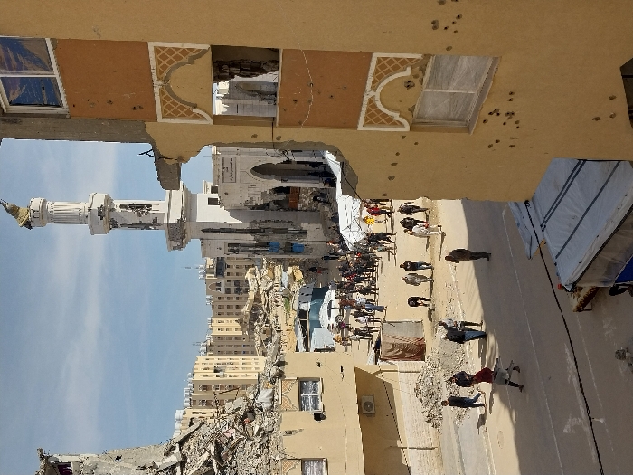 The image depicts a devastated urban street scene in Khan Yunis, Palestinian Territory, under a partly cloudy sky. In the foreground on the right, a multi-story, light-tan building shows extensive damage: a large section of its upper floors is missing, revealing exposed structural rebar. The remaining exterior wall is pocked with numerous impact marks, consistent with bullet or shrapnel damage, concentrated around a lower-floor window covered with a translucent material. An ornate, light-colored arched decorative element is visible above a missing window opening on this structure.
On the left side of the foreground, another building is largely reduced to rubble, with concrete fragments and twisted rebar visible. A paved street, marked with faint white lines and covered with scattered debris, occupies the lower and mid-ground. At the bottom, a partially obscured temporary structure with a white tarp roof is present.
Approximately 50 to 70 individuals, primarily adults in civilian clothing, are dispersed across the street and in the midground. Most are walking, singly or in small groups. One individual in the left midground wears a red top and carries a long, slender object. Another person in the center midground is bent over. Further up the street, a larger concentration of people is visible, along with several white tents or temporary shelters, and a white sedan parked on the street.
The background is dominated by a prominent white mosque with a tall minaret. The minaret's top section appears damaged, with a portion of its finial or cap missing. The main mosque building behind the minaret also exhibits structural damage, with visible facade deterioration and an Arabic inscription above an arched entrance. Beyond the mosque, a dense collection of multi-story, light-colored buildings fills the horizon, many of which show varying degrees of destruction, including collapsed sections and exposed interiors, contributing to the widespread appearance of urban devastation.