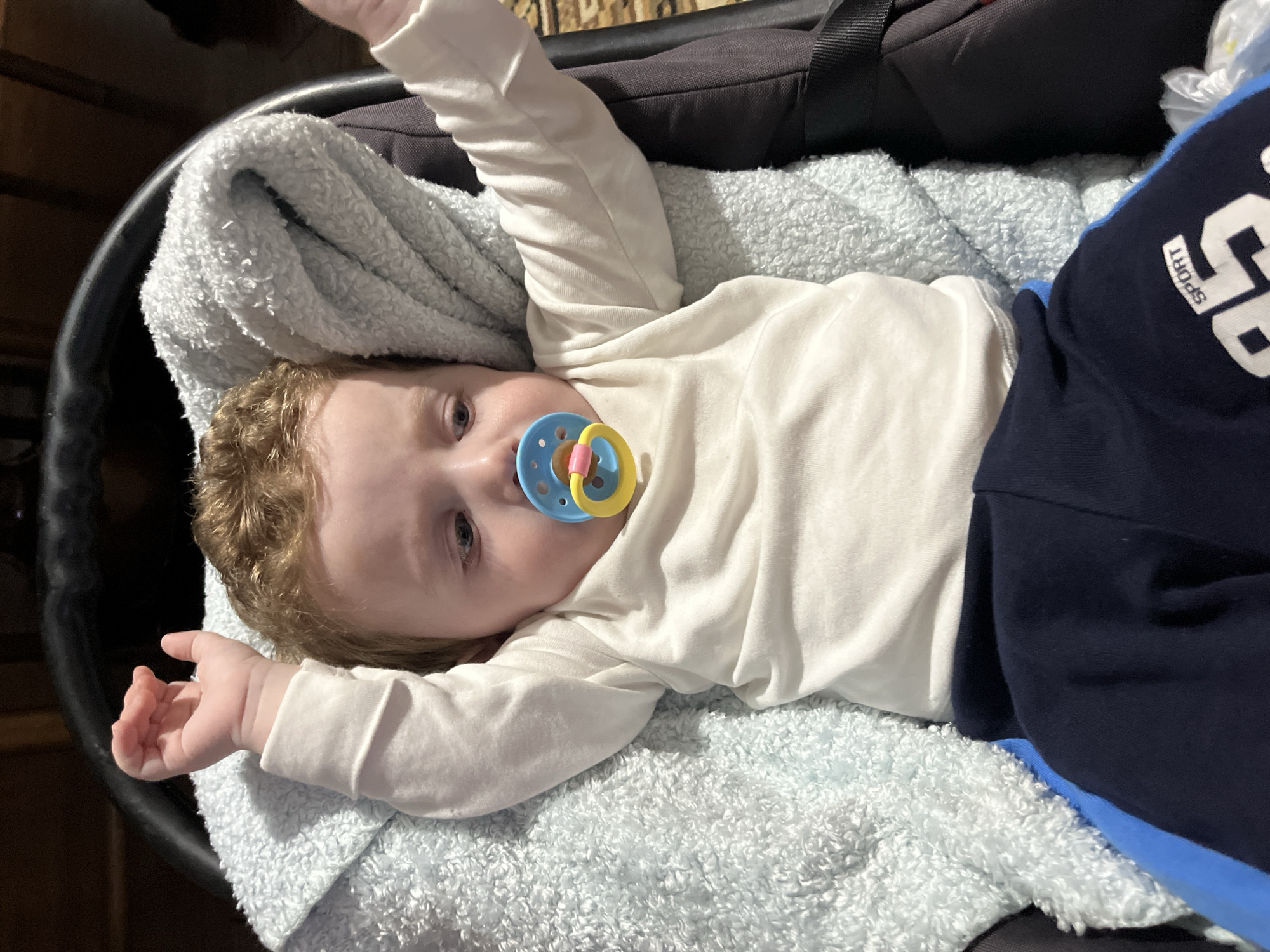 A baby with light skin and reddish-blond curly hair is lying on a light blue, textured blanket inside a black bassinet or baby carrier. The baby has a blue and yellow pacifier in its mouth. It is wearing a white, long-sleeved shirt and dark blue pants with "SPORT" and "68" printed in white on the leg. The baby's arms are outstretched. Part of a black bag is visible in the upper right corner. The setting appears to be indoors. There is no evidence of a location in Kizlyar, Russia.