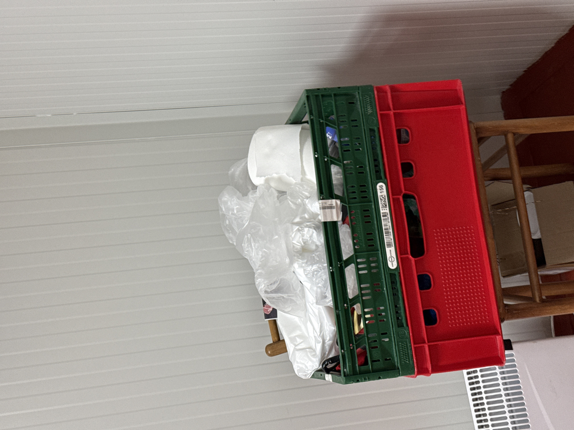 An indoor, eye-level shot captures two plastic storage crates, a green one stacked atop a red one, resting on a wooden chair or shelf. The green crate contains a roll of white paper (likely toilet paper or paper towels) and crumpled translucent plastic bags. A white label with a barcode and the number "2224156" is visible on the side of the green crate. The background features a light gray or white slatted wall or ceiling. In the bottom right corner, a white louvered grate, possibly a vent or part of a radiator cover, is partially visible beneath the red crate. The scene depicts a functional storage arrangement within an interior space in Stara Zagora, Bulgaria. No people are present, and no specific activity is taking place beyond the static arrangement of objects. The lighting suggests an indoor setting, likely under artificial light.