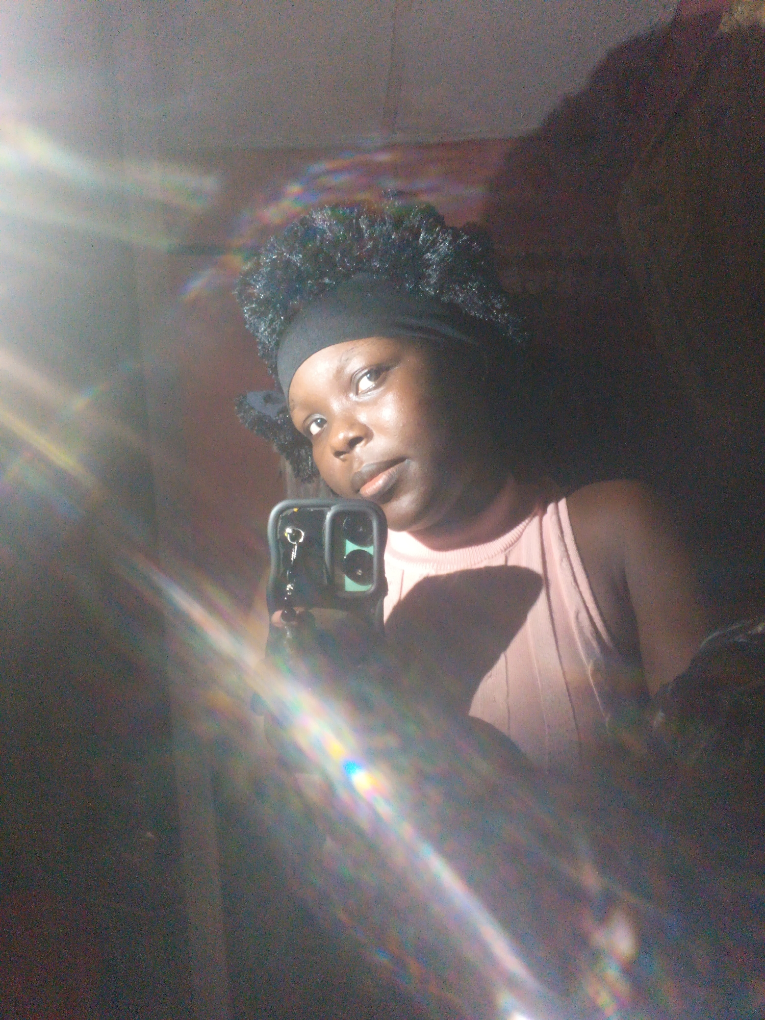 A young Black woman with curly hair styled in a black headband takes a mirror selfie. She is wearing a light pink, sleeveless top. She is holding a smartphone in her right hand, which is visible in the reflection. The lighting is dramatic, with a strong light source creating lens flare and casting shadows. The background appears to be an indoor setting with textured walls.