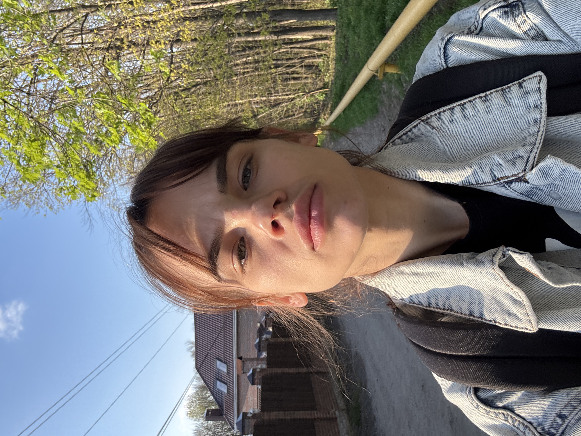 A woman with brown hair and fair skin is taking a selfie outdoors. She is wearing a denim jacket over a black shirt. A wooden railing is visible behind her. Trees and a building can be seen in the background. The location cannot be determined to be Rostov-on-Don, Russia with information available in the image.