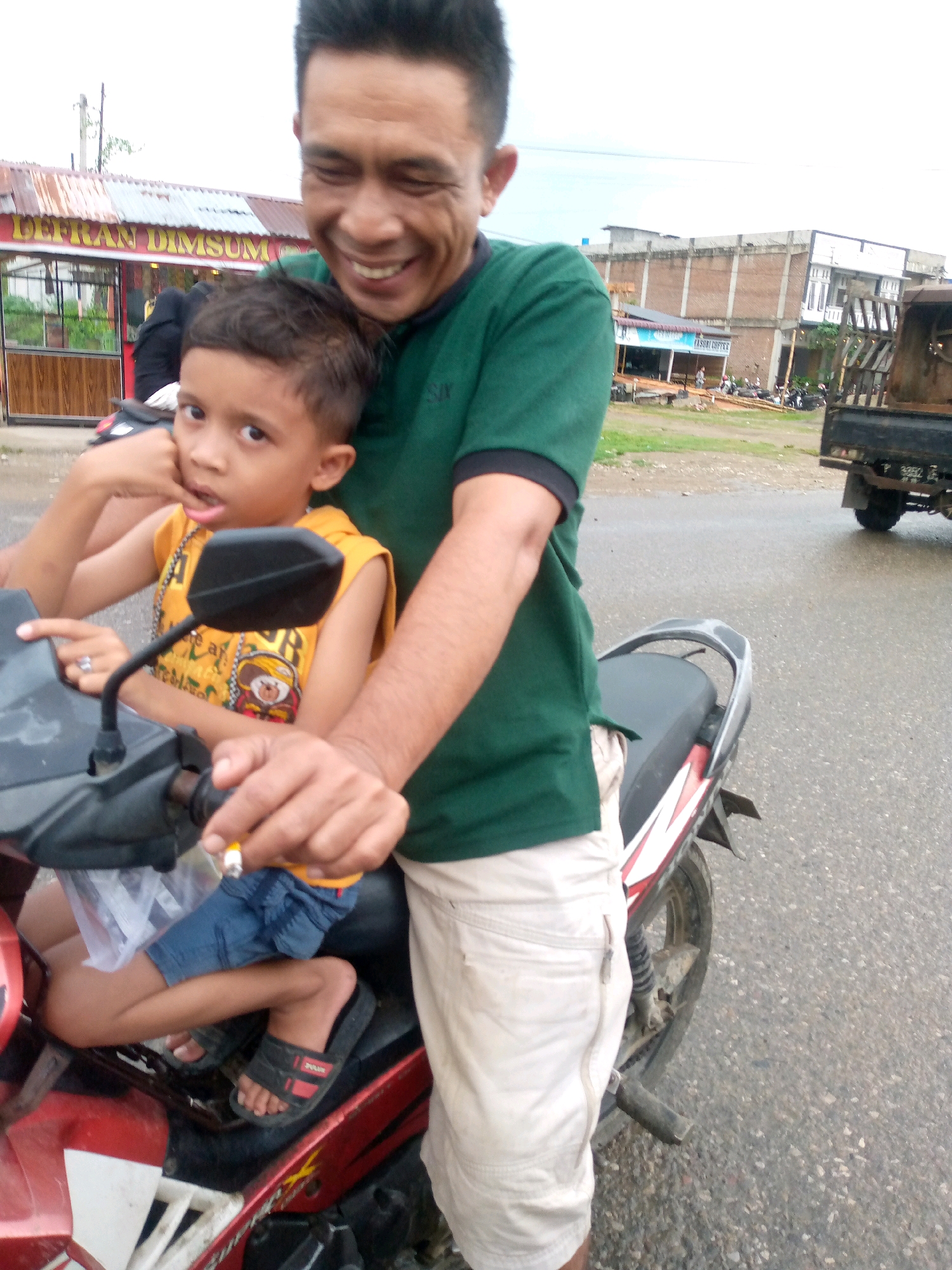 A man and a young boy are riding a red and silver motorcycle on a paved road in Gampongpaya, Indonesia. The man, with short dark hair, is wearing a green t-shirt and light-colored shorts. He is smiling broadly and holding a cigarette in his right hand. The boy, wearing a yellow tank top with a cartoon character and denim shorts, is seated in front of the man. He is holding the man's right arm with his left hand and appears to be looking at the camera with an open mouth. The motorcycle has a black seat and a prominent black rearview mirror. In the background, there are buildings, including a structure with a sign that reads "LEFRAN DIMSUM," and a truck parked on the side of the road. The sky is overcast.