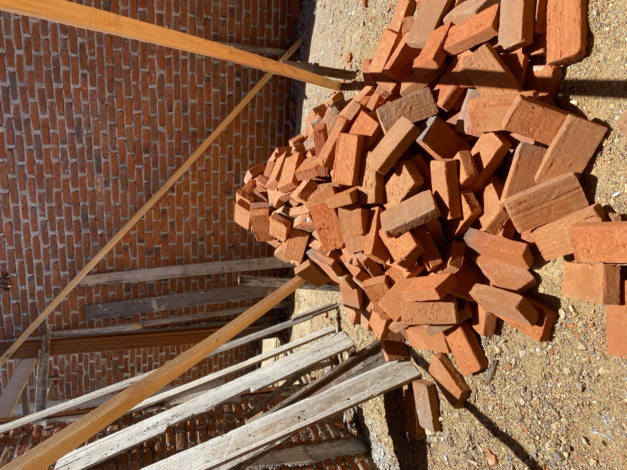 A pile of red bricks sits on a gravel surface in front of a brick wall. Wooden planks and supports are visible behind the pile of bricks and in the background. The scene appears to be a construction site.