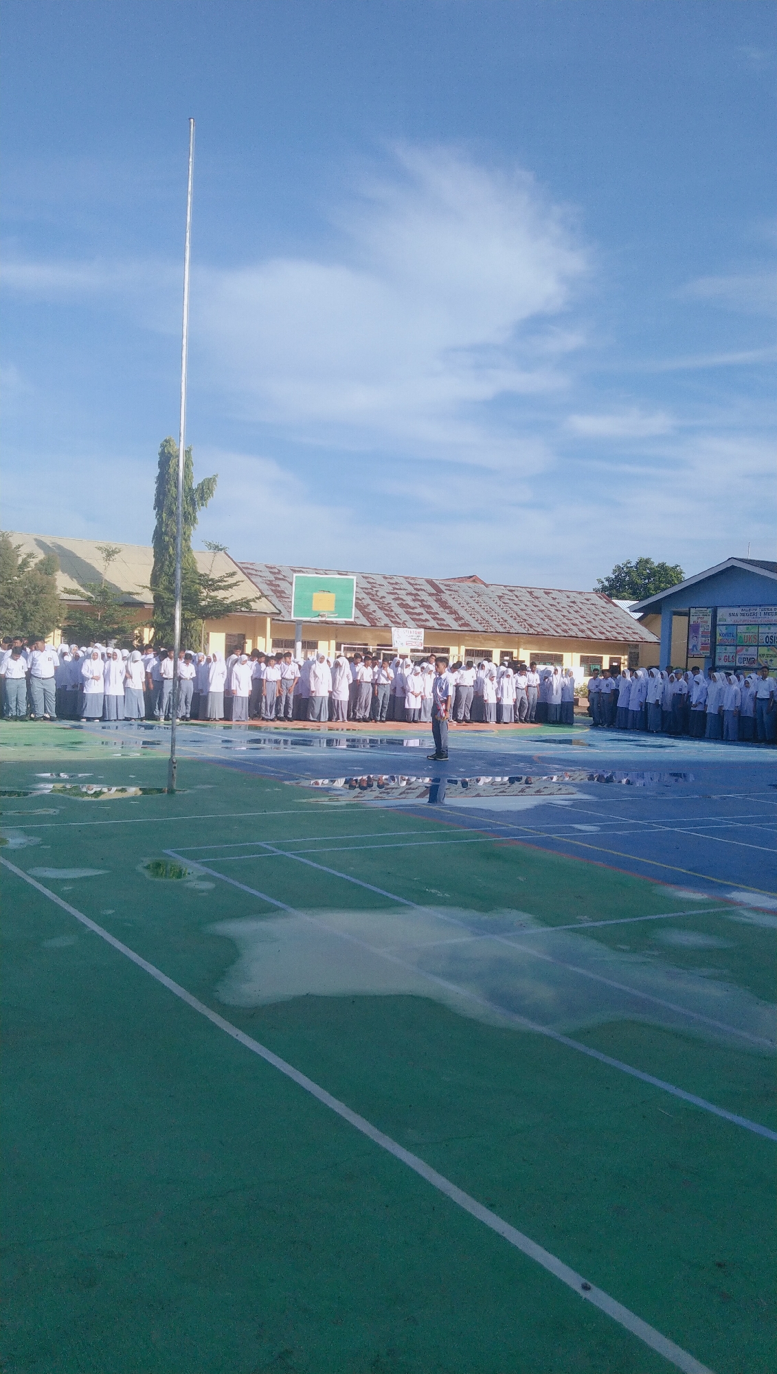 A group of students, appearing to be of high school age, stand in formation on an outdoor court. The students are dressed in white shirts and gray pants or skirts, with many wearing headscarves. They are arranged in rows, facing the camera. In the foreground, the court is a mixture of green and blue surfaces with visible water puddles, suggesting recent rain. White lines mark lanes on the court. A tall flagpole stands to the left of the frame. In the background, a building complex with a corrugated roof is visible, and a basketball hoop is mounted on one of the structures. The sky is blue with scattered white clouds. The image appears to be taken at the SMA Negeri 1 Meukek school, located in the Rhieng Blang village of the Blangpidie district, Southwest Aceh Regency, Aceh Province, Indonesia.