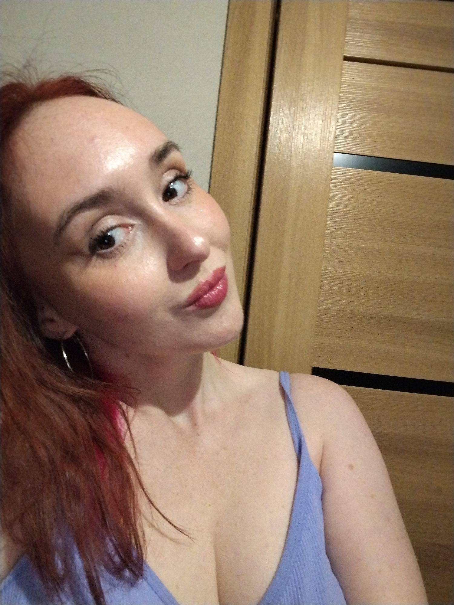 A selfie of a light-skinned woman with red hair pulled back in a ponytail. She has freckles and is wearing a light purple tank top and hoop earrings. She is looking off to the side with a slight smile. Behind her is a light-colored wooden door with black horizontal accents. There is no information present in the image to indicate that it was taken in the city of Izhevsk, Russia.