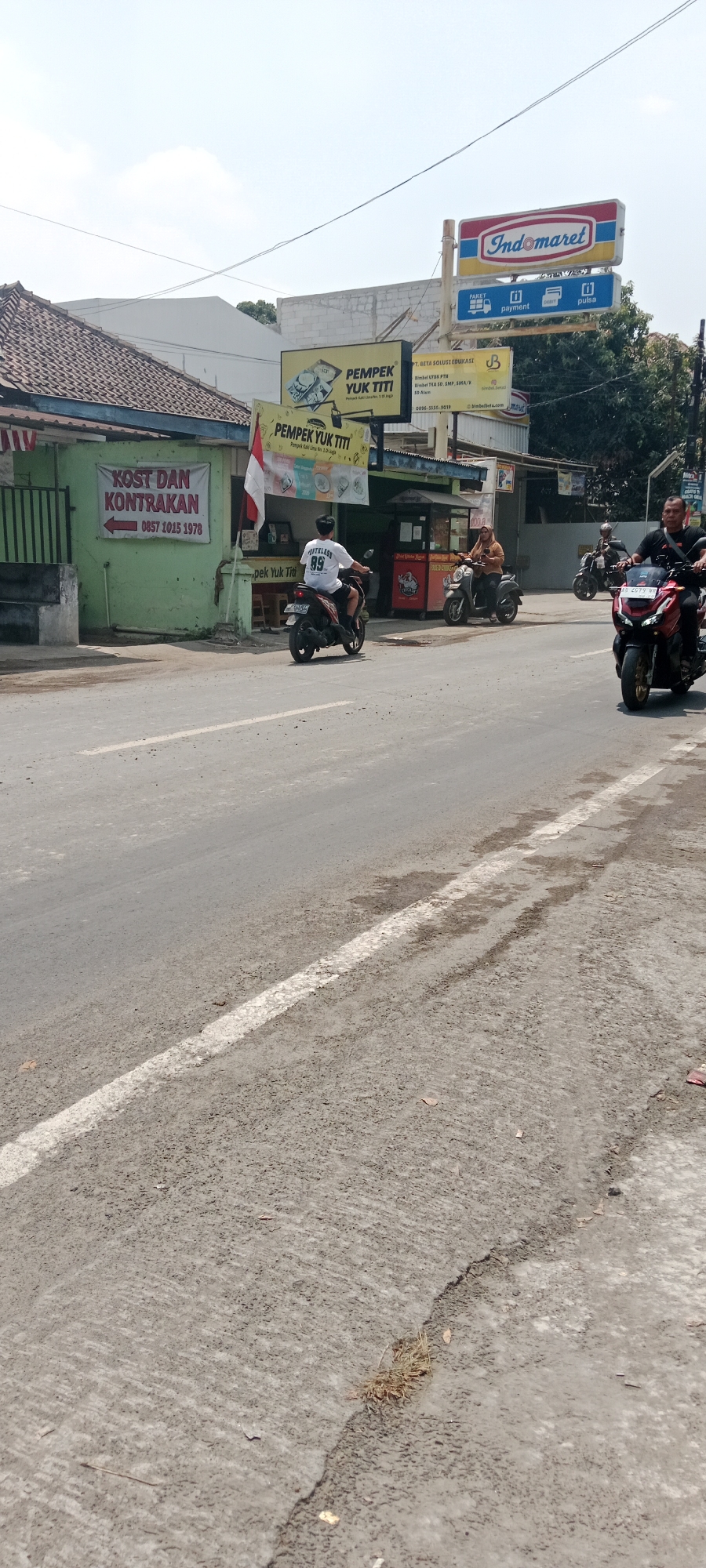 The image depicts a street scene in Maguwoharjo, Indonesia. In the foreground, a gray asphalt road with a white line marking is visible, showing some wear and tear with patches of dirt and debris. Several motorcycles are present. On the left side of the road, a green building displays a banner that reads "KOST DAN KONTRAKAN" with a phone number. A person on a red motorcycle is riding past this building. Further down the street, a person on a dark-colored motorcycle is also riding. To the right, a red motorcycle with two occupants is approaching, with the driver wearing a black shirt and the passenger in a black helmet. In the background, an Indomaret convenience store sign is prominently displayed, along with other signs advertising "PEMPEK YUK TITI." The sky is clear and bright.