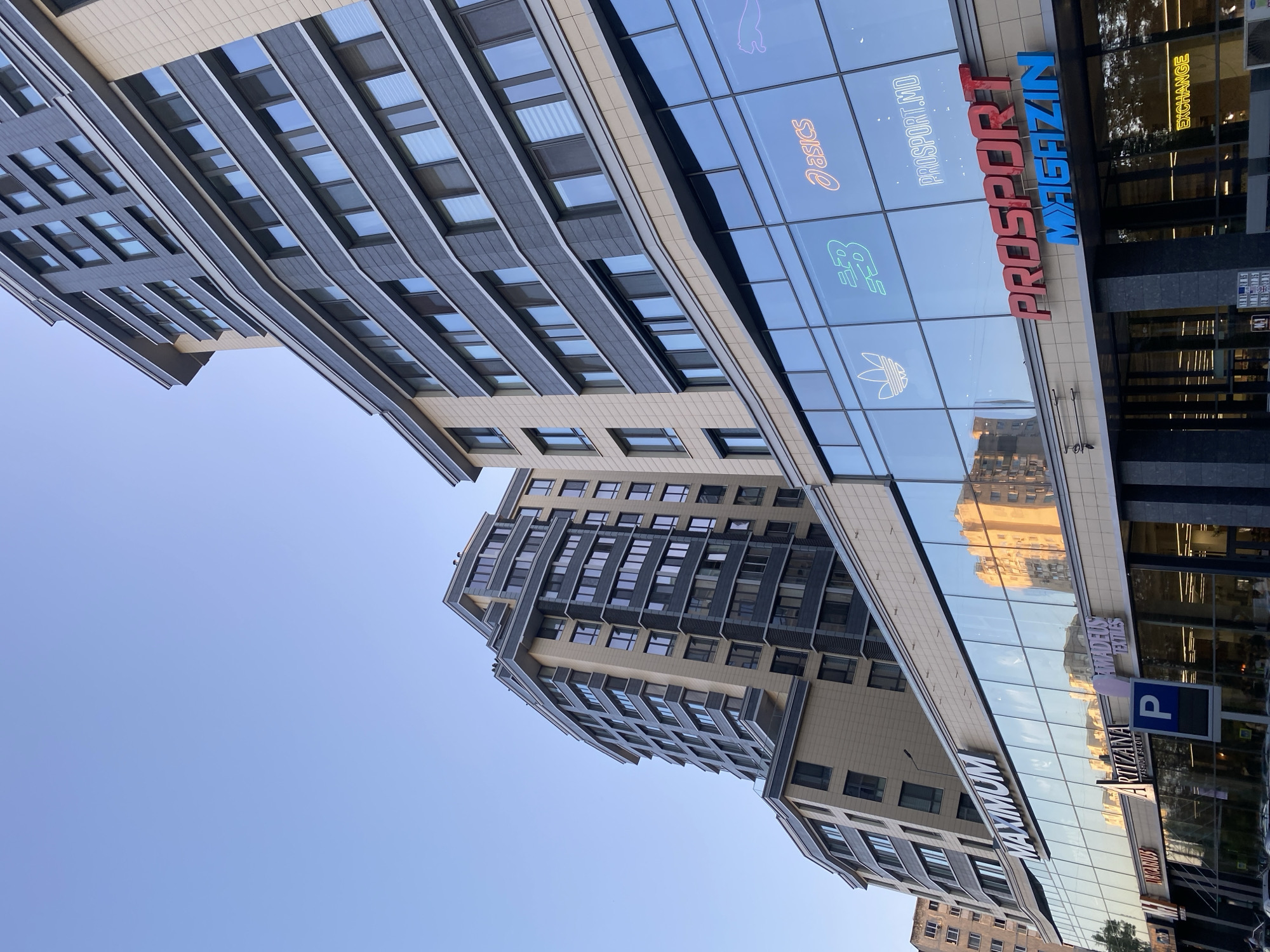The image captures a modern architectural scene in Chișinău, Moldova, likely a commercial district. The foreground features a multi-story building with a large glass facade, reflecting the sky and surrounding structures. Prominently displayed on the glass are illuminated brand logos of sporting goods companies, including Adidas, New Balance, and Asics, alongside text that reads "PROSPORT.MD" and "PROSPORT MAGAZIN". Below the glass facade, a street-level entrance to a store is visible, with signage for "MAXIMUM" above. Other visible businesses on the ground level include "ARTIZANA FASHION SALON", "AMADEUS TEXTILES", and "EXCHANGE". The background is dominated by the upper sections of several contemporary residential or office buildings, characterized by repeating patterns of windows and balconies, with a clear blue sky above. The scene suggests a well-developed urban environment with a focus on retail and modern construction.