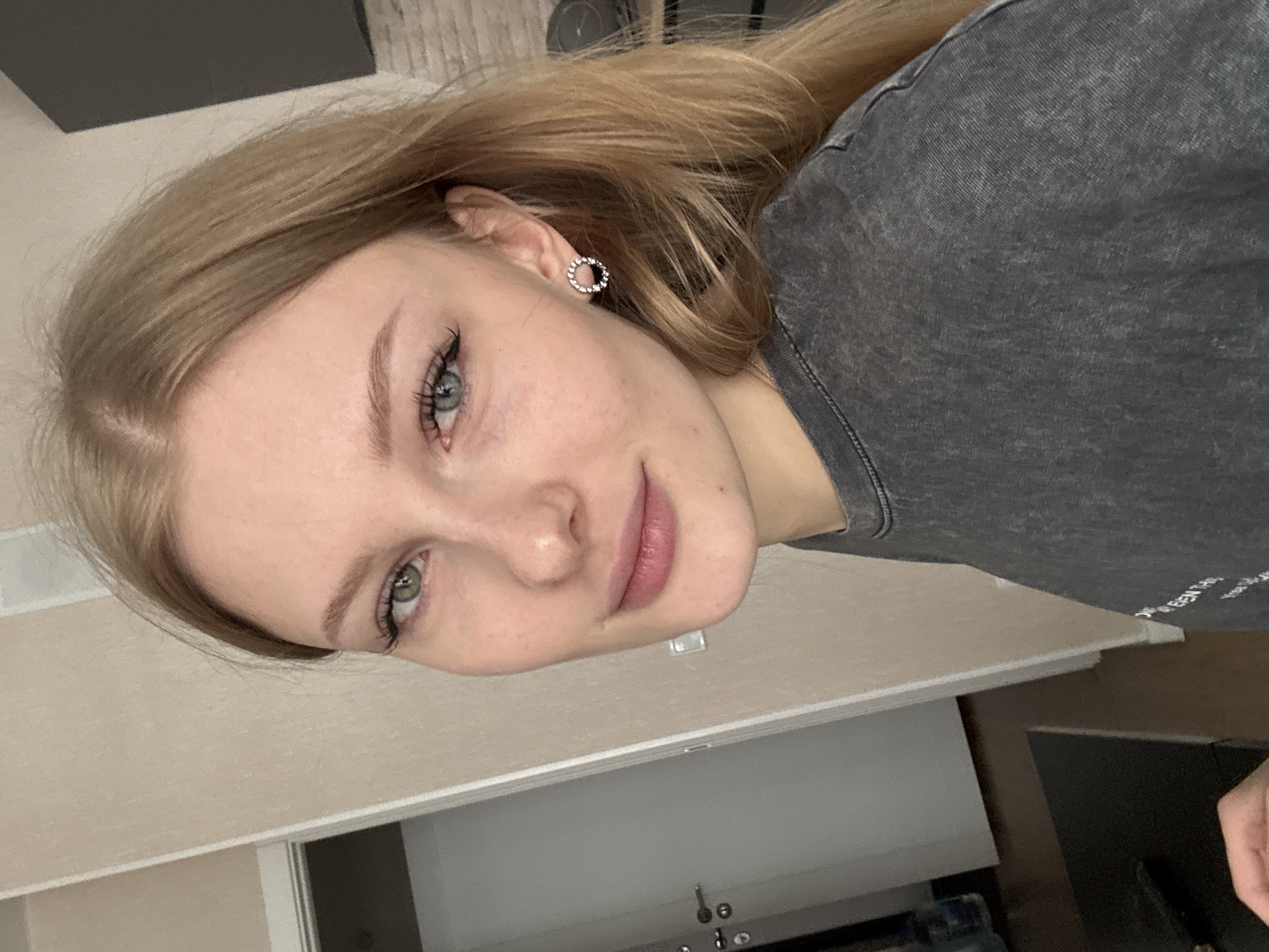 A young woman with light blonde hair, fair skin, and green eyes is seen from a close-up, head-and-shoulders perspective. She wears a dark gray t-shirt and round, silver earrings. The background shows a light-colored wall, a cabinet, and an indistinct dark object. The location is potentially a residential interior in Perm, Russia, but no specific landmarks confirm this definitively.