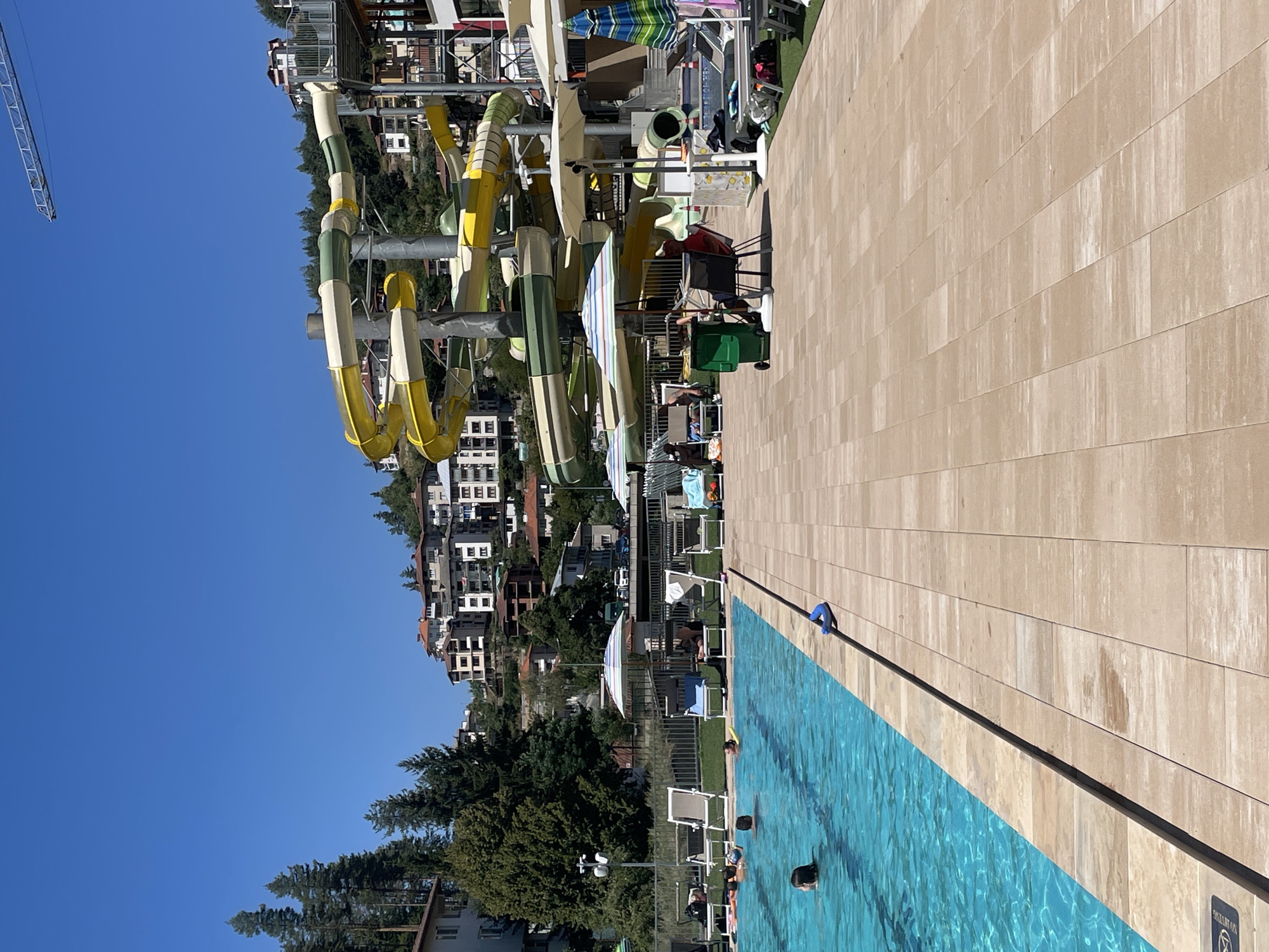 In Banite, Bulgaria, this image captures a sunny day at a water park. A large, clear blue swimming pool dominates the foreground, with a tiled deck surrounding it. Several people are in the pool, some swimming, one playing with a ball, and others relaxing. In the background, a multi-story water slide complex with yellow and green tubes is visible, a common feature in such recreational facilities. Beyond the water park, a hillside is dotted with residential buildings and lush green trees, typical of the Bulgarian landscape. The sky is a clear, bright blue, indicating fair weather.