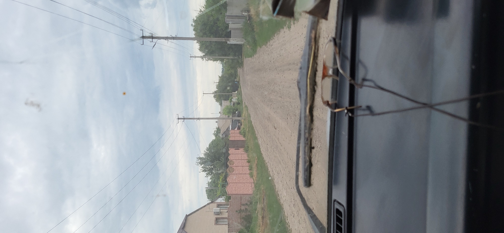 The image shows a view from inside a vehicle looking through the windshield. The vehicle is on a dirt road in a rural area, likely a village. To the left of the road, there is a house with a light-colored facade and a pink decorative fence. Further down the road, on both sides, are several houses and trees. Overhead, power lines are strung between utility poles. The sky is overcast. In the foreground, resting on the dashboard, is a pair of eyeglasses. The location is identified as Hetmanivka, Ukraine.