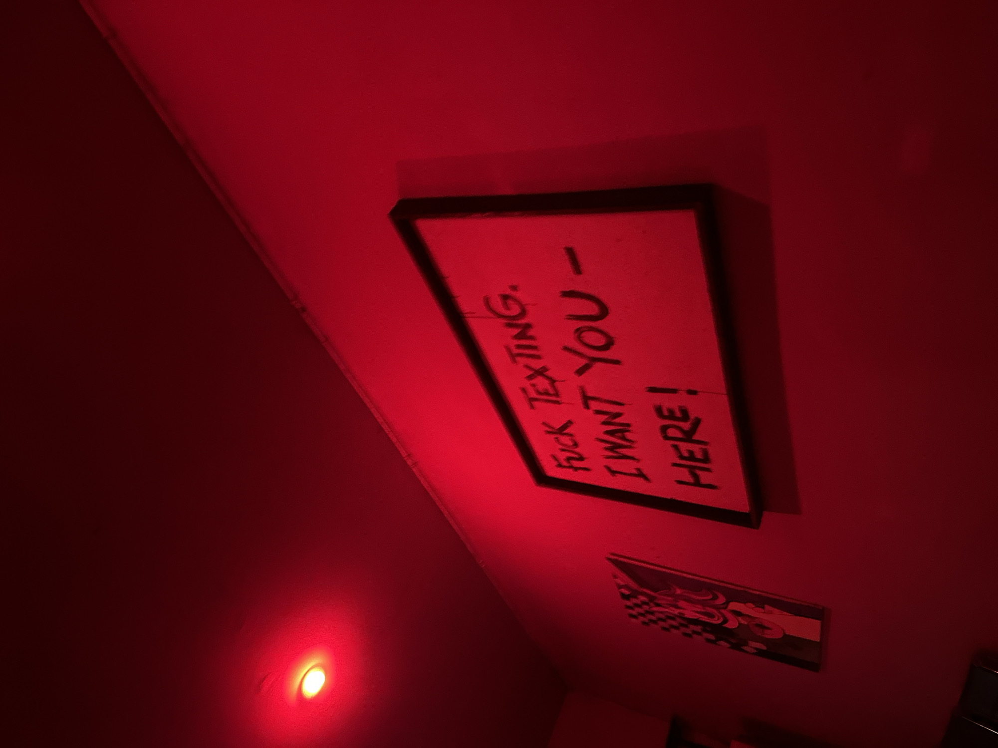 The image, bathed in red light, depicts a room's interior. On the wall hangs a framed sign with black lettering stating "F*CK TEXTING. I WANT YOU HERE!" Below and slightly to the left, another framed piece featuring a design with checkered elements and what appears to be a human-like figure. A light source casts a glow on the wall, illuminating the scene in a red hue. Based on available data, this image has been sourced from North Kuta, Indonesia. No people are visible, and details regarding the environment beyond the immediate visible interior are unavailable.