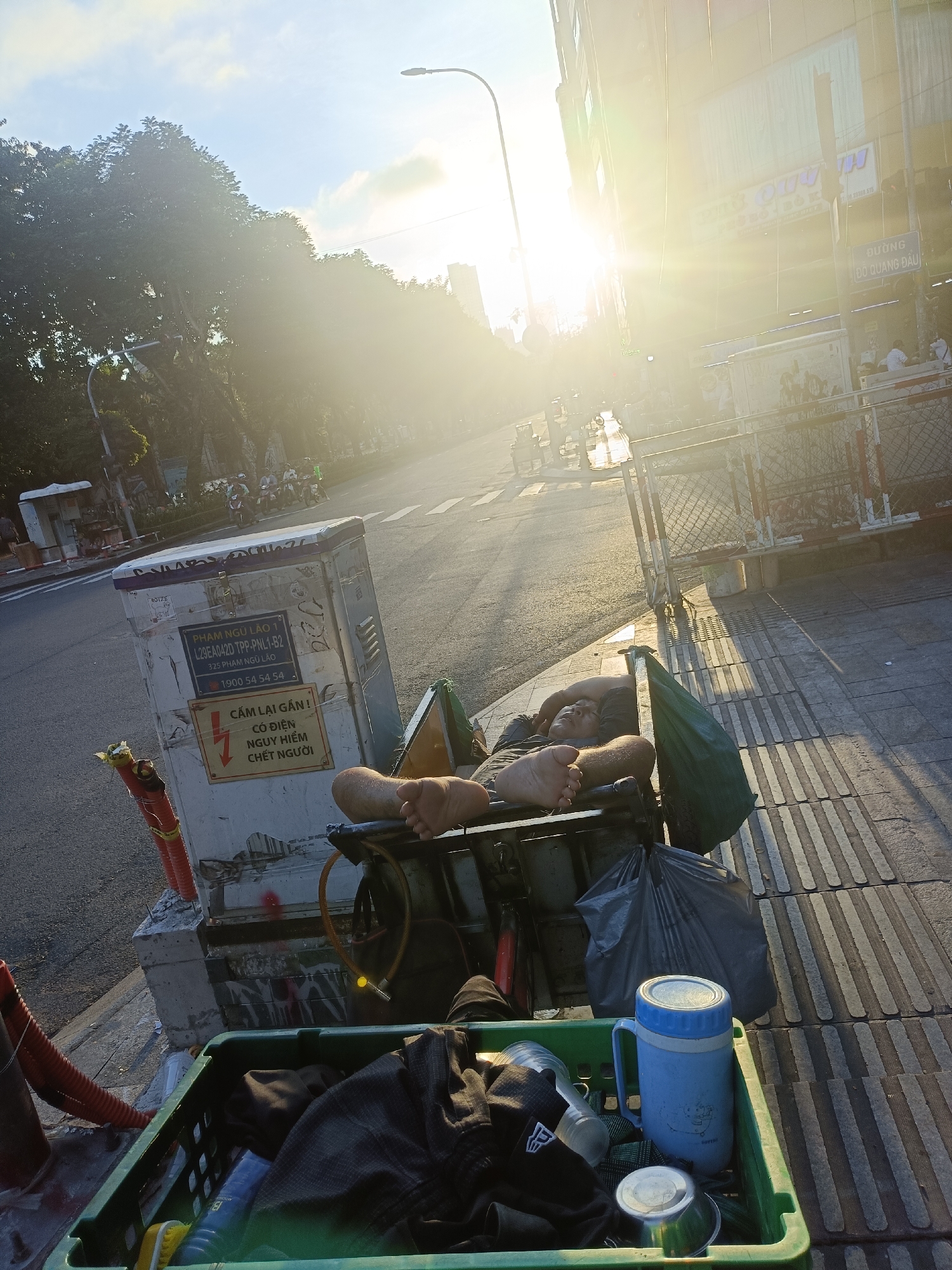 In Thủ Đức City, Vietnam, a person is sleeping in a makeshift bed constructed on a cart next to a utility box on a sidewalk. The cart is covered with a green tarp and has a large black trash bag and a blue thermos. A green plastic crate filled with clothing and other items is also present. The utility box has labels and warnings, including a lightning bolt symbol indicating a danger. The scene is illuminated by bright, low-angle sunlight, suggesting it is either early morning or late afternoon. In the background, there is a street with traffic, including motorbikes, and buildings. Trees line the street.
