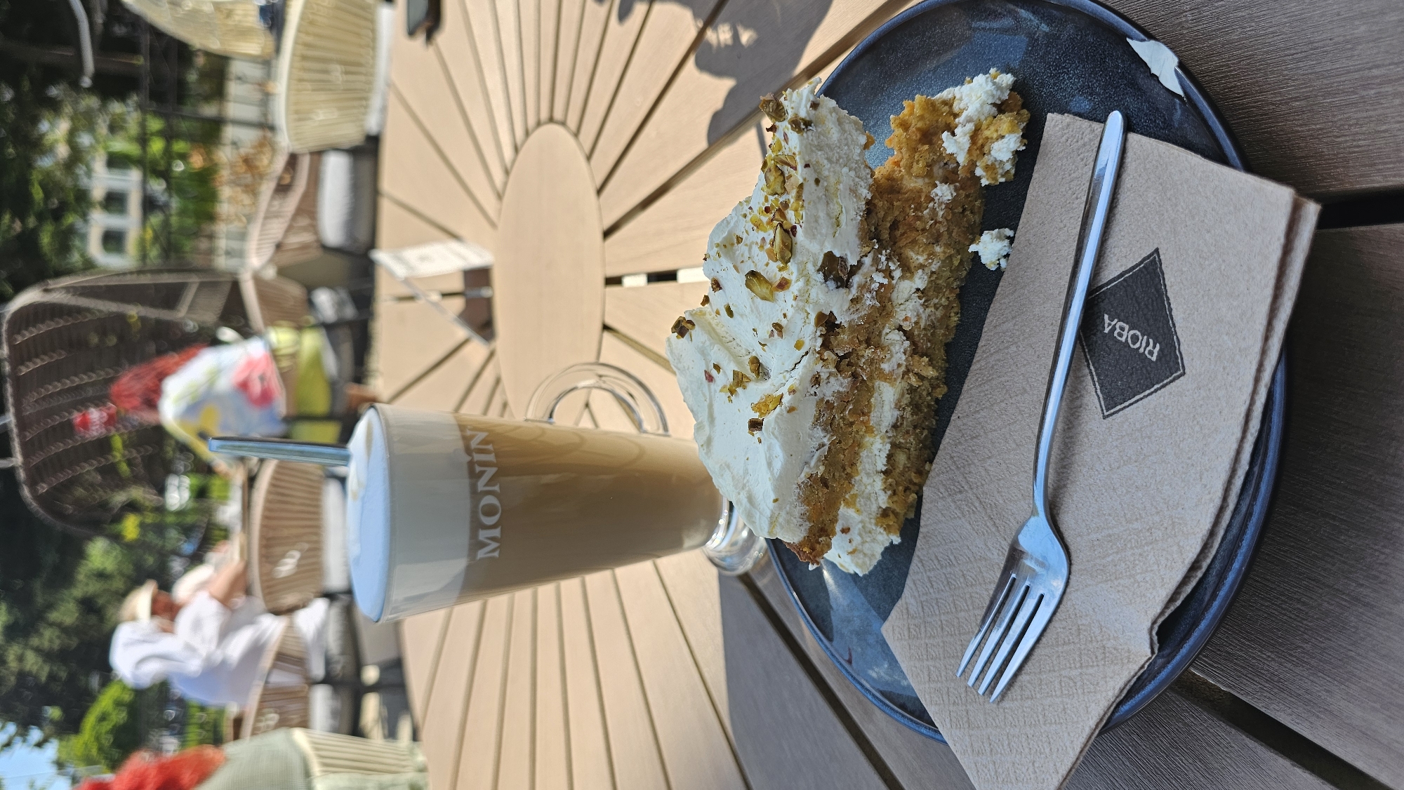 The image shows an outdoor cafe table. A slice of cake with white frosting and pistachios sits on a dark blue plate, next to a folded brown napkin with the word "RIOA" printed on it. A silver fork rests on the napkin. A glass of latte or coffee with a white froth top is also present. The table is made of light-colored wood. People are visible in the blurry background, including a person in a white shirt and hat, and chairs arranged around tables. The image was taken in Varna, Bulgaria.