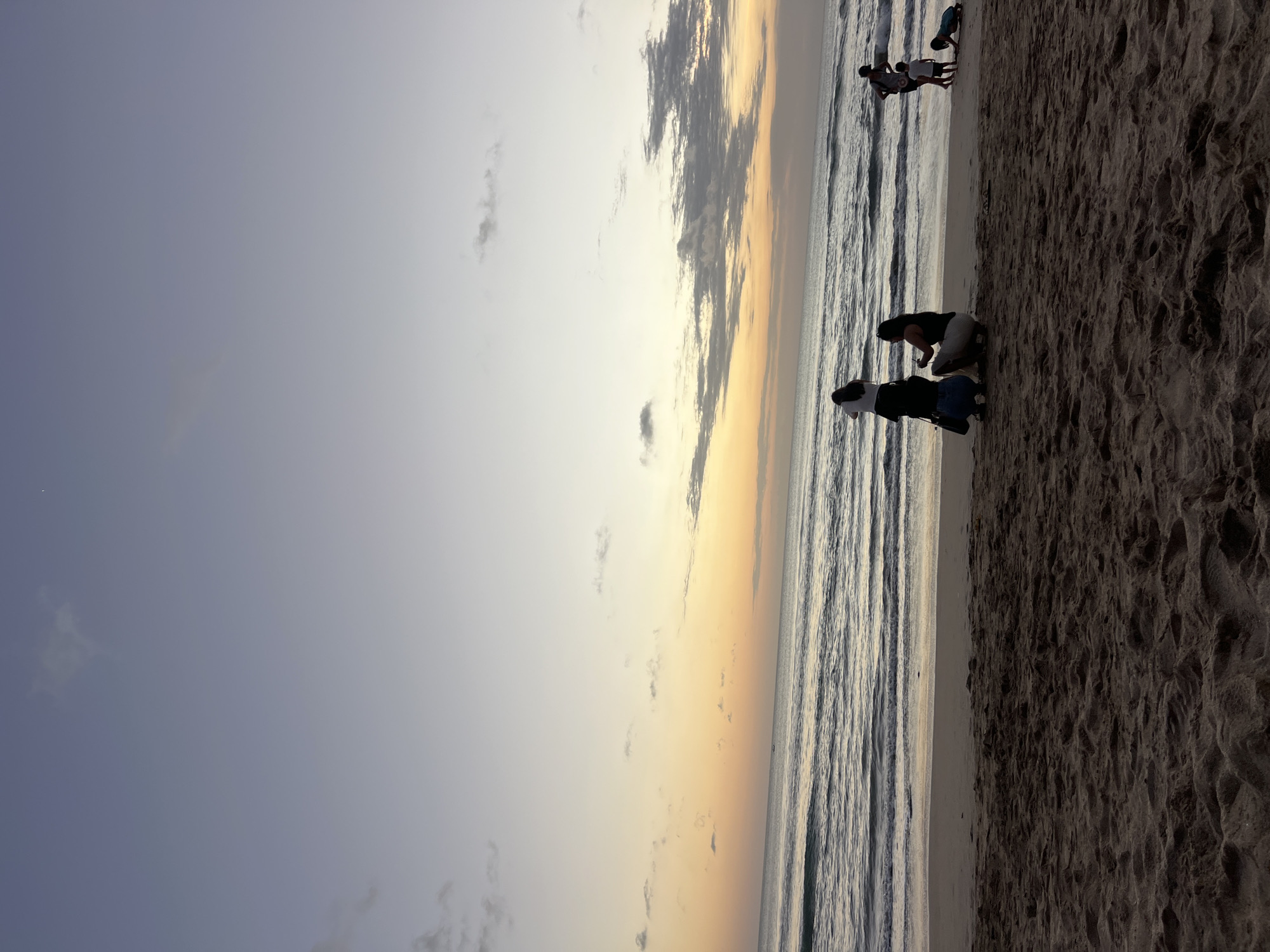 The image depicts a beach scene in Kuta, Indonesia, likely during sunset or twilight. The sky transitions from a pale blue at the top to a warm orange and yellow hue near the horizon. Gentle waves roll onto a sandy shore, which is textured with footprints and uneven sand. Several groups of people are scattered across the beach. In the foreground, two individuals are kneeling on the sand, one with a white shirt and dark pants, the other in a dark tank top and light skirt. Further down the beach, a group of four people, including two adults and two children, are visible near the water's edge. One child is crawling on the sand, while others appear to be interacting. The overall atmosphere suggests a peaceful evening at a popular tourist destination.