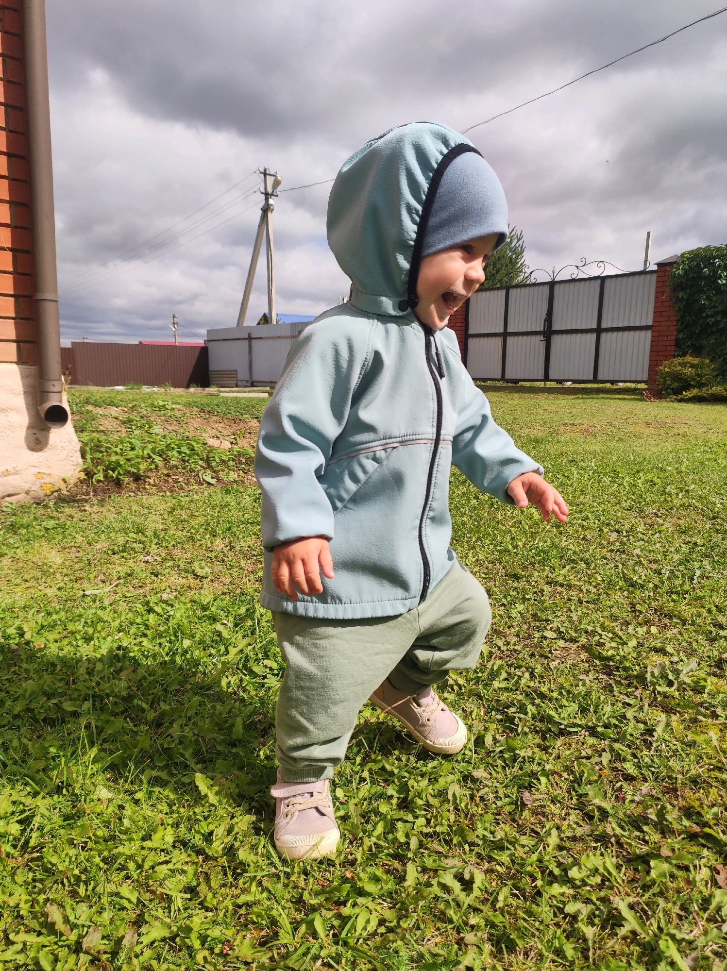An outdoor medium shot captures a young child from the waist down in a playful stance on a grassy lawn under a cloudy sky. The child, wearing a light blue hooded jacket, a blue beanie, and green pants, is looking to the right with their mouth open in a laugh. The child's right hand is extended forward. The background includes a brick building with a downspout on the left, a wooden fence, a metal gate, and a power line pole. The setting appears to be a residential yard in the city of Fokino, Russia.