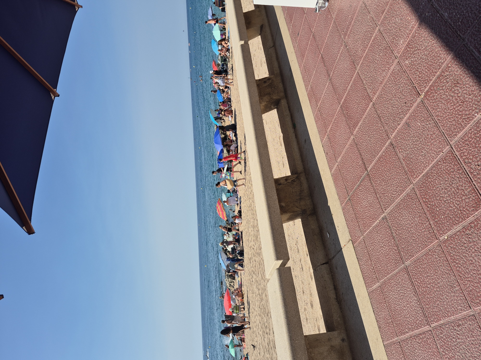 The image depicts a busy beach scene on a sunny day in Lloret de Mar, Spain. In the foreground, a red tiled walkway leads towards a sandy beach. A concrete bench separates the walkway from the beach. The beach is populated with numerous people relaxing under colorful umbrellas. Some individuals are wading in the ocean, while others are sitting or standing on the sand. The blue sea stretches to the horizon, dotted with a few distant boats. A large, dark blue umbrella is partially visible in the upper portion of the image, casting a shadow. The overall impression is one of a vibrant, lively beach resort.