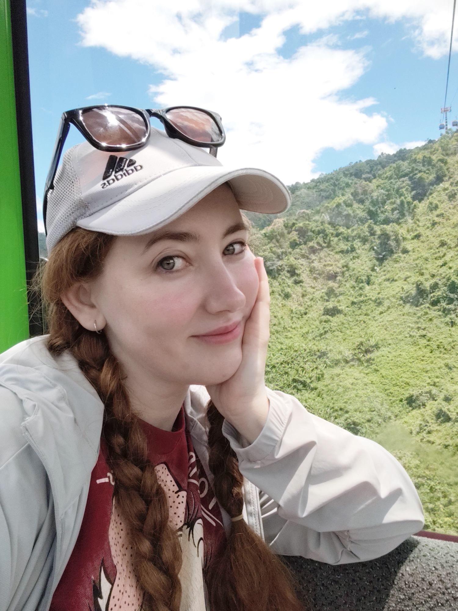 A young woman with reddish-brown hair in two braids is taking a selfie in a cable car, looking at the camera with a gentle smile. She is wearing a light grey jacket over a red t-shirt with a graphic print and a white baseball cap with sunglasses resting on top. The background shows a lush green mountainside and a blue sky with scattered clouds, and a glimpse of a cable car system further up the mountain. The cable car appears to be moving through a mountainous, forested area.