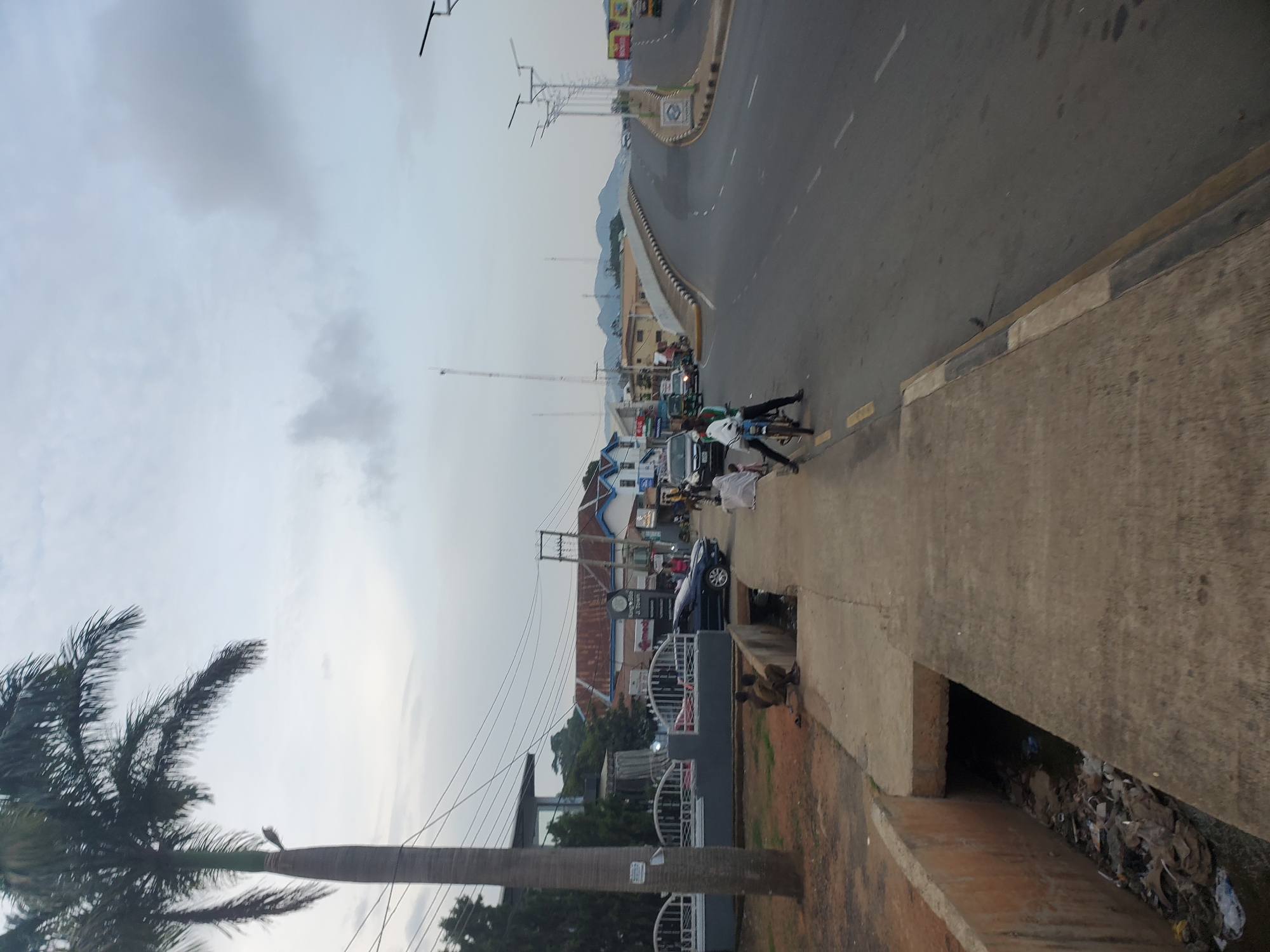 A wide-angle, eye-level outdoor shot captures a street scene in Jos, Nigeria, under an overcast sky. In the foreground, a concrete sidewalk runs along the left side of the frame. A shallow, concrete-lined drainage ditch, filled with debris and trash, runs along the edge of the sidewalk. To the left of the sidewalk, a large palm tree stands tall, its fronds reaching towards the sky. Beyond the sidewalk, several cars are parked and driving on a paved road. Buildings with colorful signs line the street. In the midground, a modern building with a glass facade is visible. Further back, a traffic bridge curves over the road, with streetlights lining its edge. The background shows a hazy view of hills and mountains. Several people are present in the scene, some walking, others on bicycles or motorcycles. Two individuals are seated near the drainage ditch. The overall impression is one of a bustling urban environment, likely a commercial or residential area within Jos.