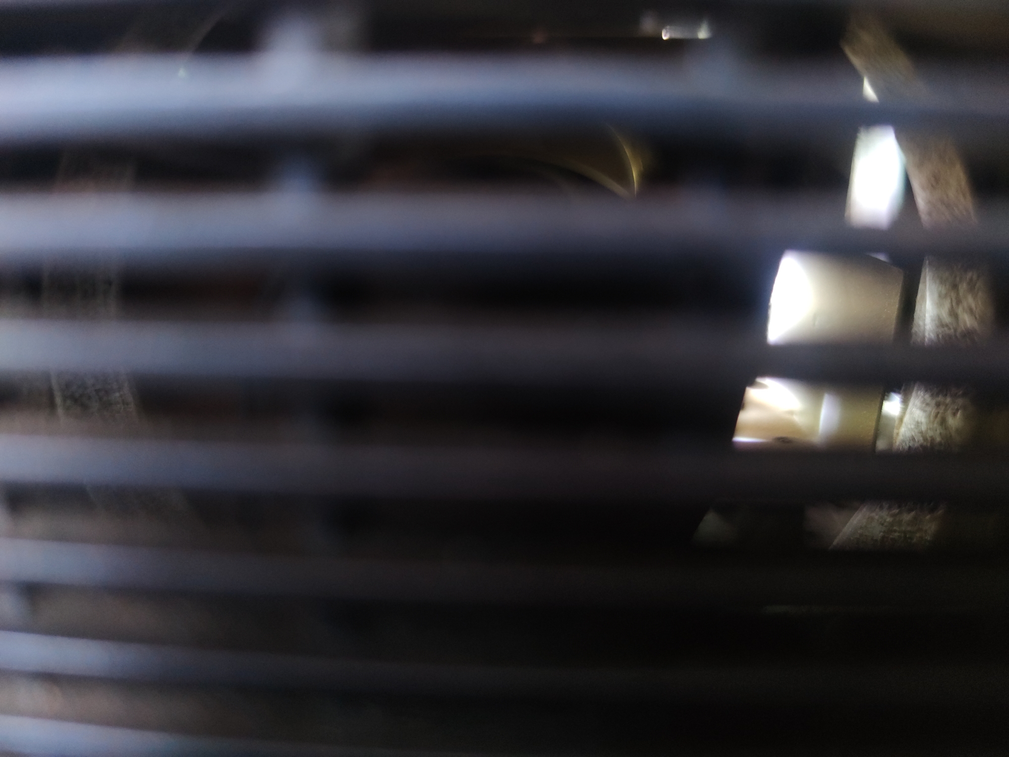 The image shows a view through horizontal slats, possibly window blinds or ventilation grates. The slats are dark and out of focus. Beyond the slats, there is a glimpse of a light-colored interior, possibly a room, with some indistinct objects or shapes visible. The specific location and identifiable subjects are not discernible from the image.