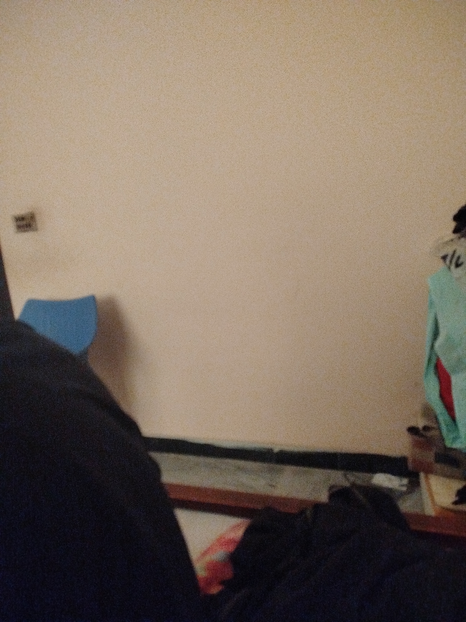 The image shows a room interior with a plain, textured beige wall that occupies most of the frame. In the lower left, a dark blue fabric is partially visible, appearing to be clothing or bedding. To the right of this, a bright blue object, possibly a cushion or part of a piece of furniture, casts a shadow on the wall. In the upper left corner of the wall, a small, dark, rectangular object is mounted. On the right side of the image, a light blue garment, possibly a shirt or jacket, hangs on something out of frame, with a hint of red fabric underneath. A wooden frame runs horizontally across the lower portion of the image, suggesting the presence of a bed or furniture. The floor area below the wall appears to be a dark surface. The scene is dimly lit and appears to be a private living space. No specific location in Lassan, Pakistan can be identified from the visual information.